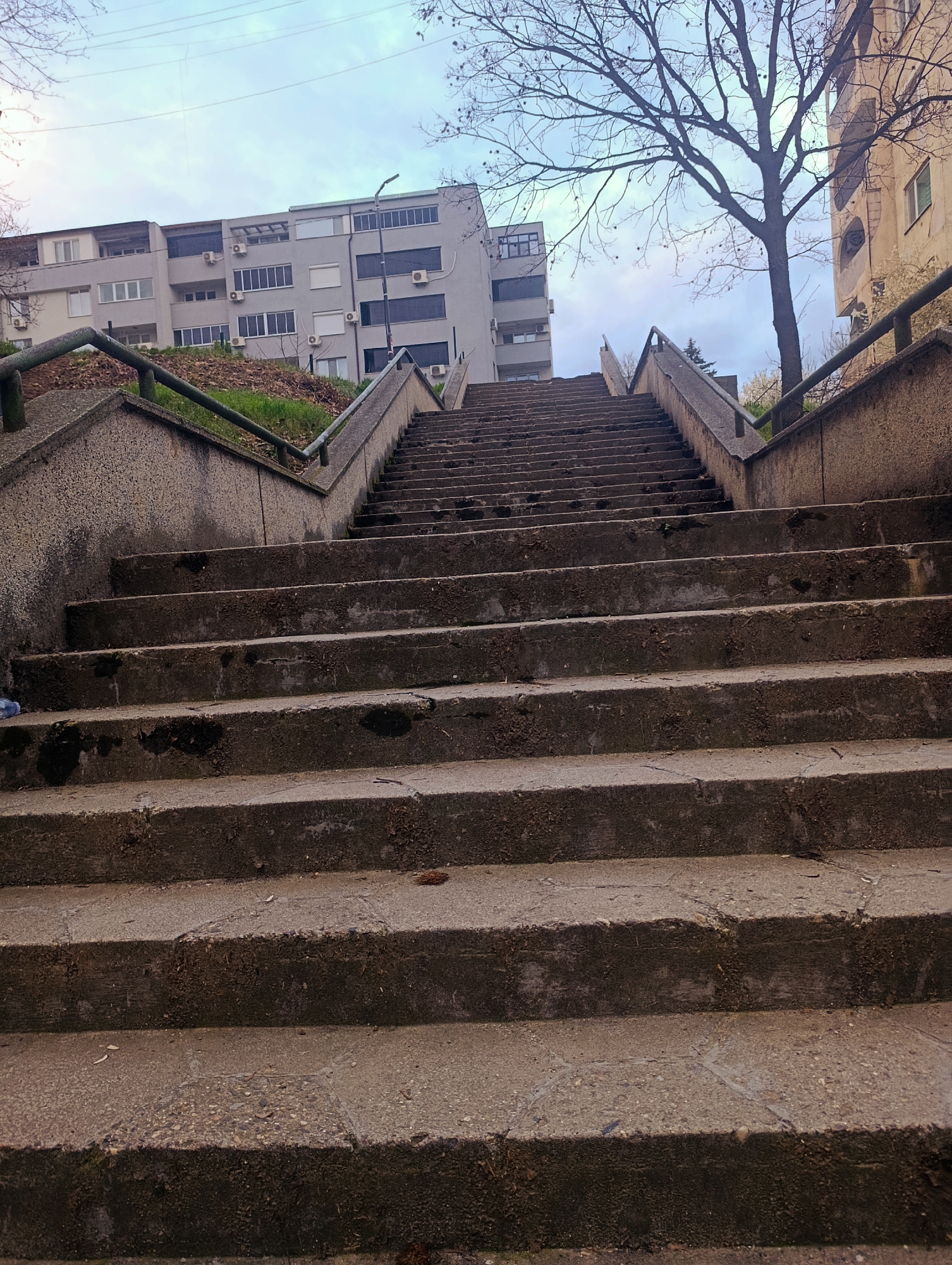 The image depicts a long, weathered concrete staircase leading uphill. The stairs are flanked by low concrete walls topped with metal handrails. Several buildings are visible in the background, including an apartment building with multiple windows and what appears to be air conditioning units. A bare tree stands to the right of the staircase. The sky is overcast. The location is the stairs near the Perperikon Hotel in Kardzhali, Bulgaria.