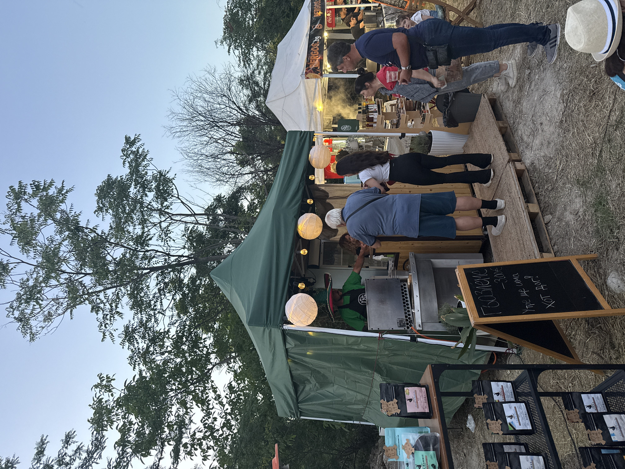 The image depicts an outdoor market scene, likely in Stara Zagora, Bulgaria, with a focus on a coffee stall. A green tent is set up, with a vendor wearing a green shirt and a distinctive hat serving customers. Bags of coffee are displayed on a shelf to the left of the tent. In front of the tent, a chalkboard sign displays prices and what appears to be a greeting.
To the right of the green tent, a white tent is also set up, branded with "BERGOS.BG" and featuring a Coca-Cola logo. This tent appears to be a food stall, with steam rising and people ordering. Several individuals are visible interacting with vendors and each other, suggesting a lively market atmosphere. The ground is a mix of dirt and straw, with trees and foliage surrounding the stalls. The sky is clear and blue, indicating daytime. A child is visible sitting in a chair near the white tent, observing the scene. The overall impression is of a community gathering or event, possibly an evening market given the lighting from the lanterns.