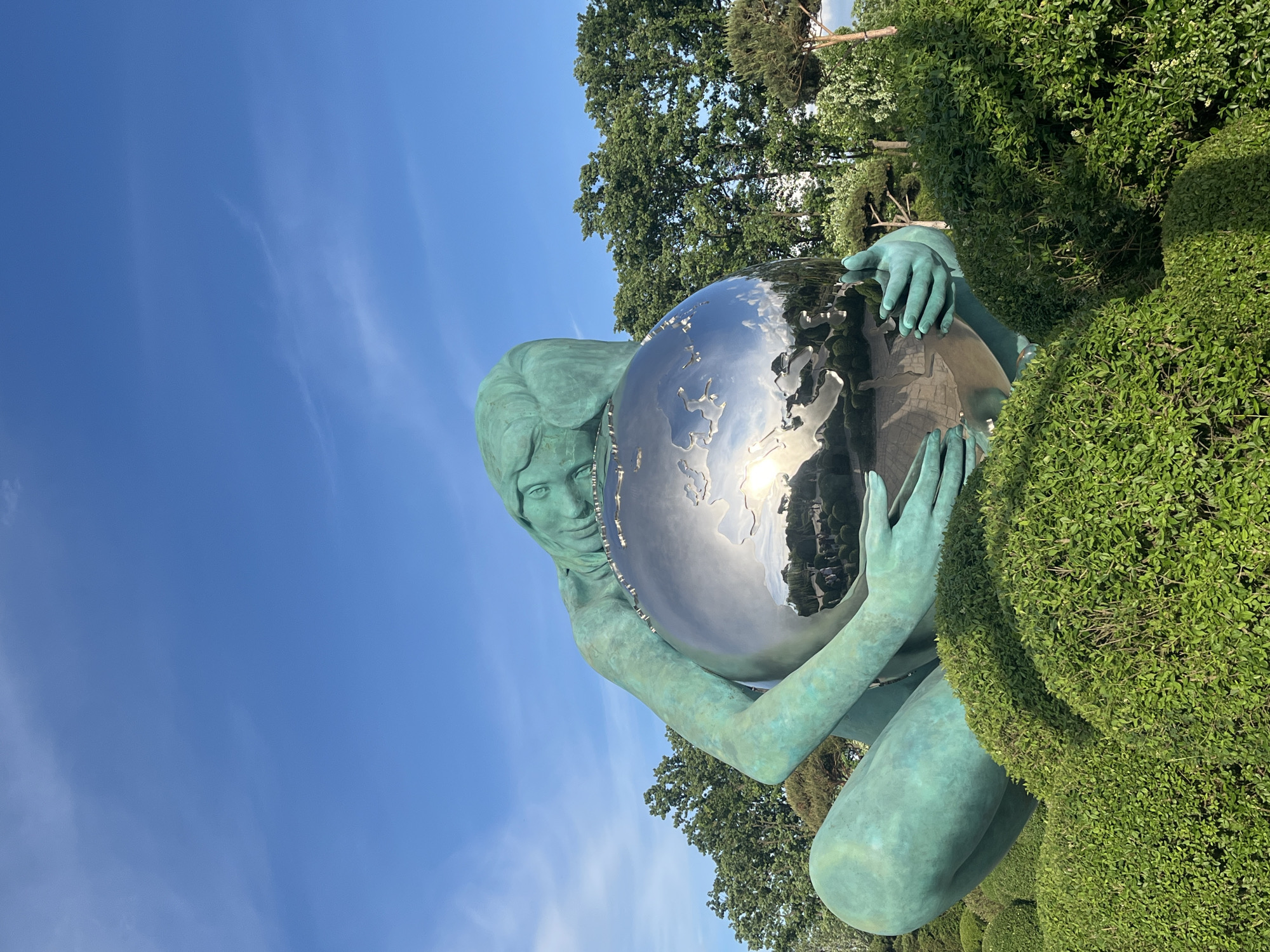 A bronze-colored statue of a woman in a squatting position holding a shiny silver globe is positioned among sculpted green hedges in a park setting. The woman is rendered with her hair swept back and a somber expression. The globe reflects the surrounding environment, including trees and sky. Blue sky with faint clouds is visible in the upper left of the frame. Green trees are present in the background. The image appears to be taken at a slight angle. This is likely a photograph of the "Globe of Peace" sculpture in Krasnodar, Russia.