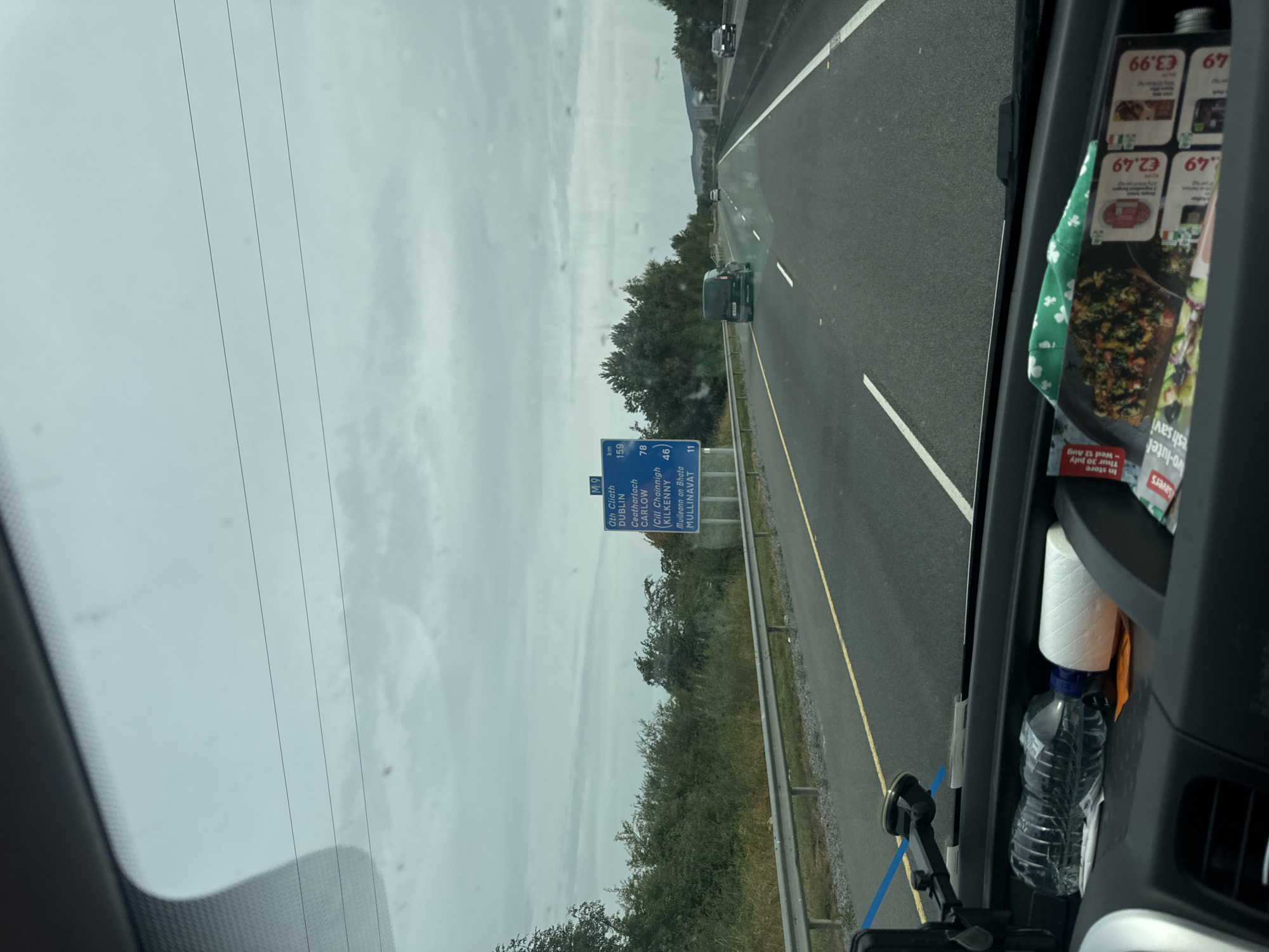 This image is a point-of-view shot from inside a vehicle, likely a van, looking forward through the windshield. The vehicle is traveling on a highway in Grannagh, Ireland, as indicated by the M9 motorway sign which lists destinations and distances, including Dublin (159 km), Carlow (78 km), and Kilkenny (46 km). The sky is overcast and grey, with visible horizontal lines across the windshield that appear to be defroster elements.
On the right side of the road, a blue directional sign displays "M9" at the top, followed by Irish place names and distances in kilometers. Below the sign, there is a roadside barrier. The highway itself has white lane markings. Several vehicles are visible on the road: a dark van in the right lane, a white car further ahead, and another dark car in the distance on the left. Dense green foliage lines the left side of the highway, with some trees visible further back.
Inside the vehicle, in the foreground, the dashboard is visible. On the dashboard, there is a clear plastic water bottle with a blue cap, a large spool of white thread, and several pieces of paper, including what appears to be a flyer with food images and prices in Euros. A phone mount with a blue strap is attached to the windshield. The overall impression is a typical highway journey under a cloudy sky.
