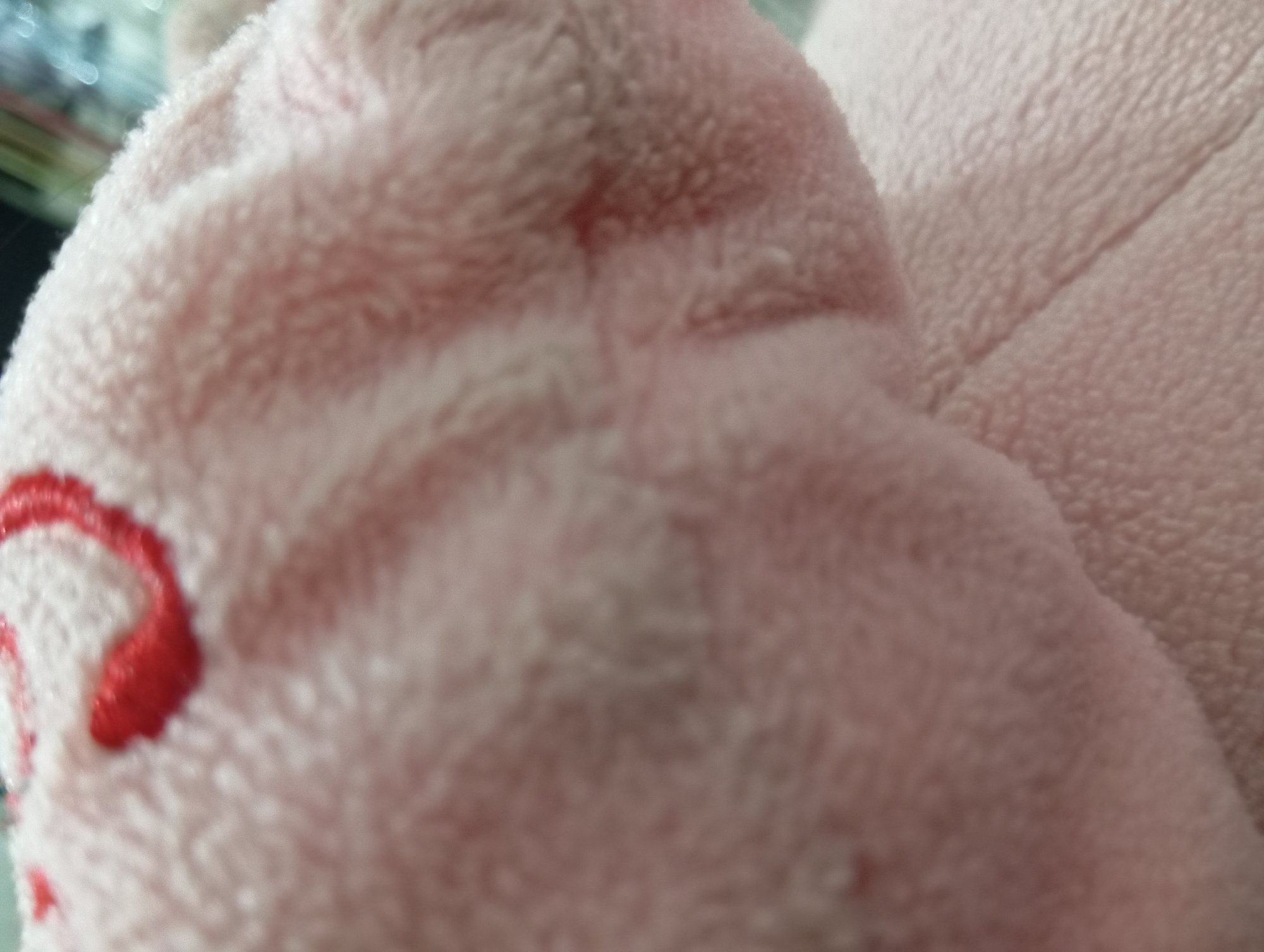 The image is a close-up, out-of-focus shot of a pink, fuzzy material, possibly a stuffed animal or blanket. There are some red embroidered or sewn details visible at the top left of the frame, forming curved shapes. The texture of the material is soft and plush, with visible fibers. The lighting is diffuse, creating soft shadows and highlights. There is no indication of the city of Rejosari, Indonesia, or any specific environmental context within the image. The scene is devoid of any people or explicit actions.