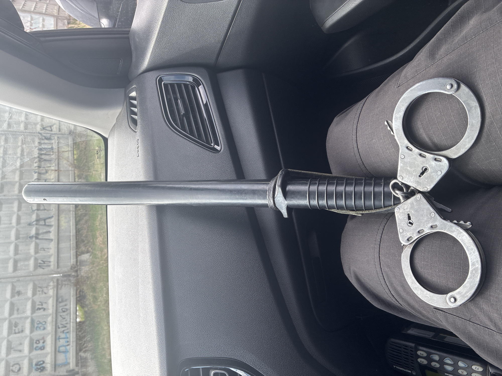 The image shows a black expandable baton and a pair of handcuffs resting on the lap of a person wearing gray pants inside a car. A Motorola radio is visible below the baton and handcuffs. The car's dashboard and vents are visible, along with a partial view out of the driver's side window showing a concrete fence with graffiti on it. The location appears to be an urban environment.