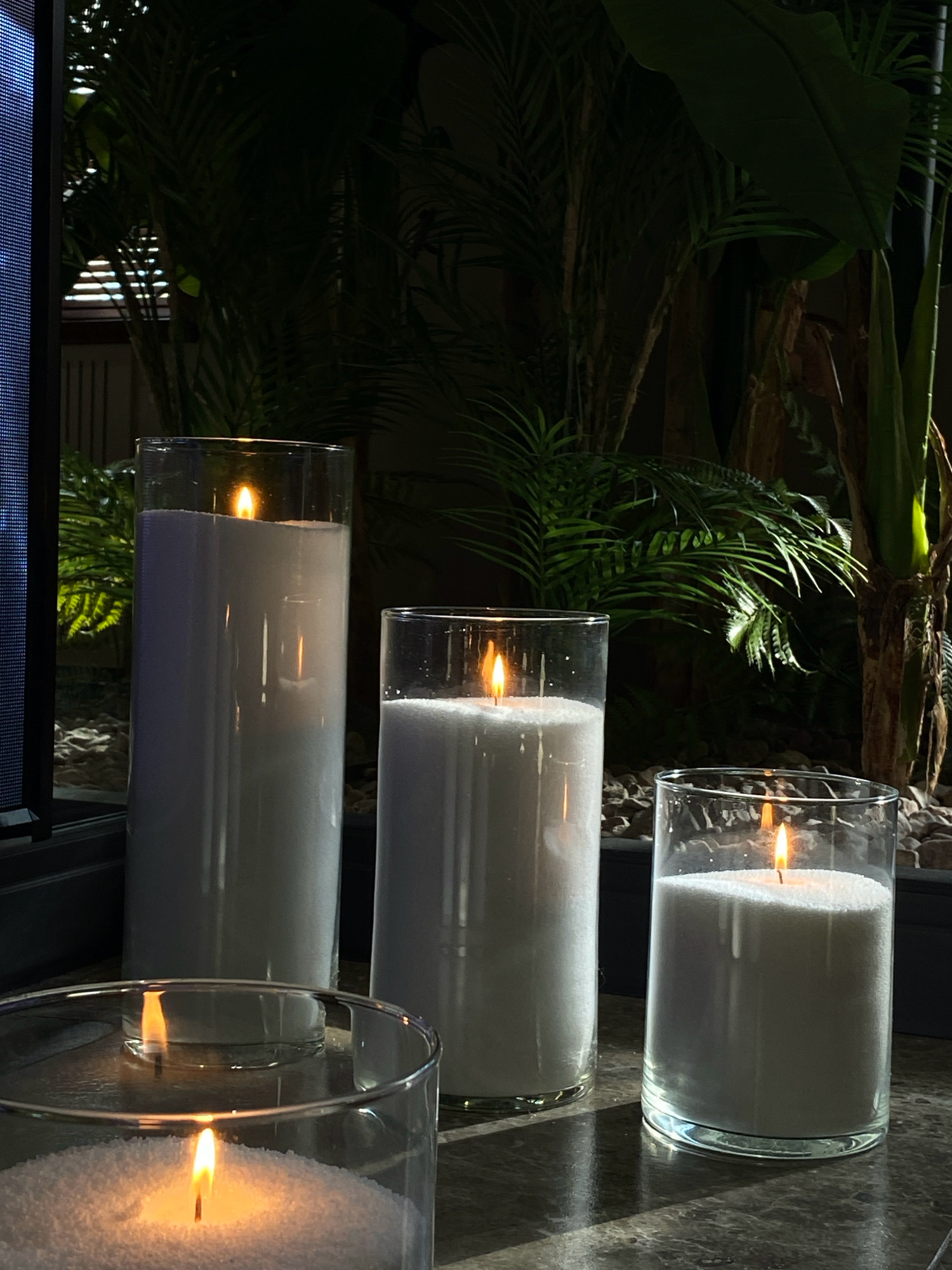 The image depicts a dimly lit interior scene featuring three lit pillar candles in clear glass cylinders filled with white granular material. The candles are of varying heights, with the tallest on the left, a medium-sized one in the center, and the smallest on the right. The flames of the candles cast a warm glow, illuminating the granular substance within and creating reflections on the surface below. In the background, lush green foliage, likely houseplants, is visible, some of which are illuminated by shafts of sunlight. The environment appears to be indoors, possibly near a window or a glass door, given the presence of a dark frame on the left edge of the image, possibly part of a window screen or frame. The overall ambiance is tranquil and serene. No people are present in the image. The image does not contain any elements that would place it specifically in Votkinsk, Russia.