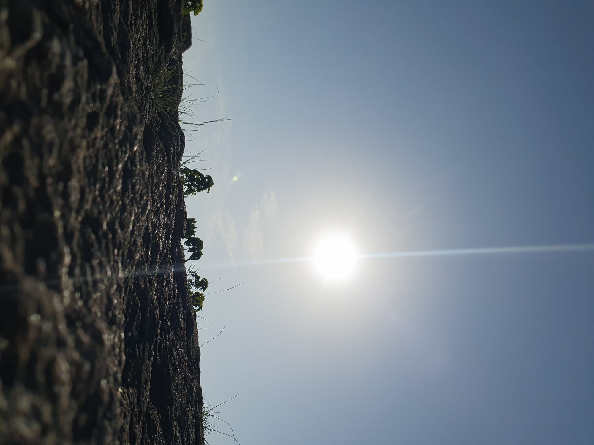 The image depicts a low-angle shot of a rocky surface under a clear blue sky with the sun prominently displayed. The sun is a bright, almost blown-out circle at the top-center of the frame, with a strong lens flare extending vertically downwards. The rocky foreground is rough and textured, with sparse blades of grass and a few small plants growing from crevices. The environment appears natural and arid, possibly a hillside or elevated terrain. The scene is devoid of any human presence or activity. The lighting suggests it is daytime, likely mid-morning or afternoon due to the sun's position and intensity. The location is not identifiable from the image alone, and no specific details suggesting Jos, Nigeria, are visible.