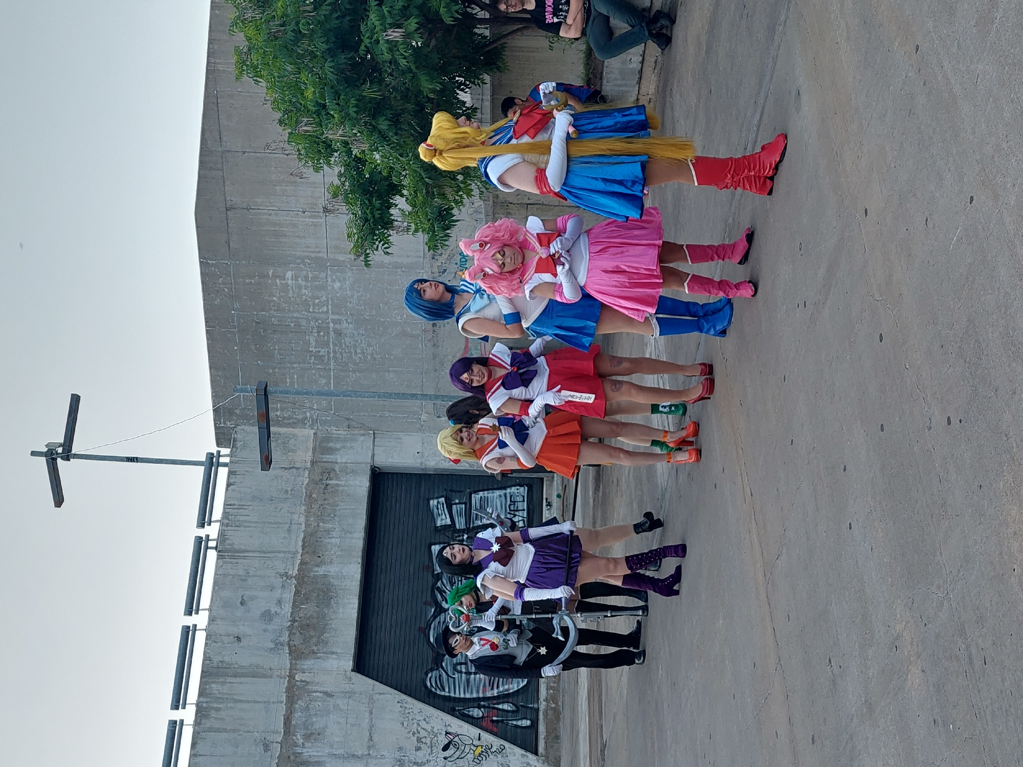 A horizontally oriented photograph captures a group of cosplayers dressed as characters from the "Sailor Moon" franchise. They are standing on a concrete surface resembling a sidewalk or street. Behind them is a gray, weathered concrete wall. To the left of the frame is a metal pole with a cross-shaped structure at the top, possibly a light pole. To the right of the pole, a large gray metal door with graffiti on the lower portion is visible.
The cosplayers are arranged in a line, from the bottom to the top of the frame, each dressed in costumes that correspond to a character from "Sailor Moon," including Sailor Moon, Sailor Venus, Sailor Mars, Sailor Jupiter, Sailor Mercury, Sailor Chibi Moon, Sailor Neptune and Tuxedo Mask. Their poses are static and the setting has minimal interaction between them, with them posing together. The location appears to be an urban, public area in Buenos Aires, Argentina.