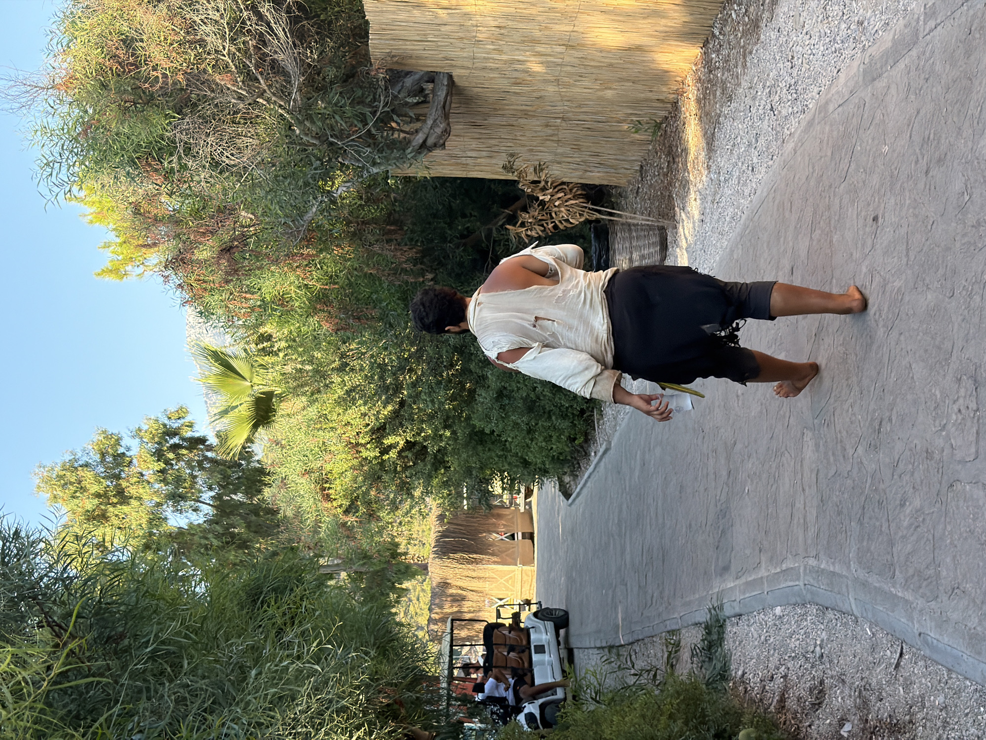 A man walks barefoot down a stone pathway. He is wearing a torn, light-colored shirt and dark, loose-fitting pants. He carries a white cup in his right hand. The path is bordered by gravel and lush green foliage, including trees and bushes. In the background, a white golf cart with passengers is visible on the left, and a structure covered in thatch or straw is on the right. The scene appears to be outdoors, possibly in a resort or garden area, given the presence of the golf cart and tropical vegetation. The lighting suggests it is daytime, with the sun casting warm light on the scene. The man's back is to the camera, and he is walking away from the viewer. The environment is consistent with a location in Fethiye, Turkey, known for its Mediterranean climate and coastal resorts.