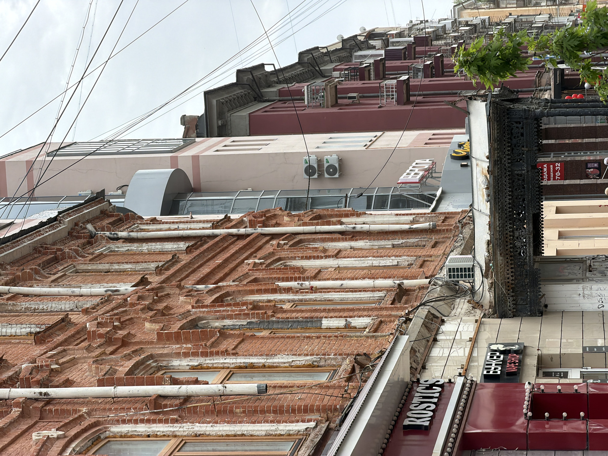 The image shows a low-angle view of several buildings in Saratov, Russia. The nearest building features a complex, multi-tiered brick facade with pipes running along it and rectangular windows. A sign reading "ROSTIC'S" is visible on its side, along with the sign of a "BUNKER VAPE SHOP". The adjacent building has an ornate black metal structure and tiled exterior. The building above the first one has a beige section with twin air conditioners attached to it. Above that is a red building with a terraced roof and multiple AC condensers. Above the buildings, electrical wires cut across the sky. The sky is overcast.
