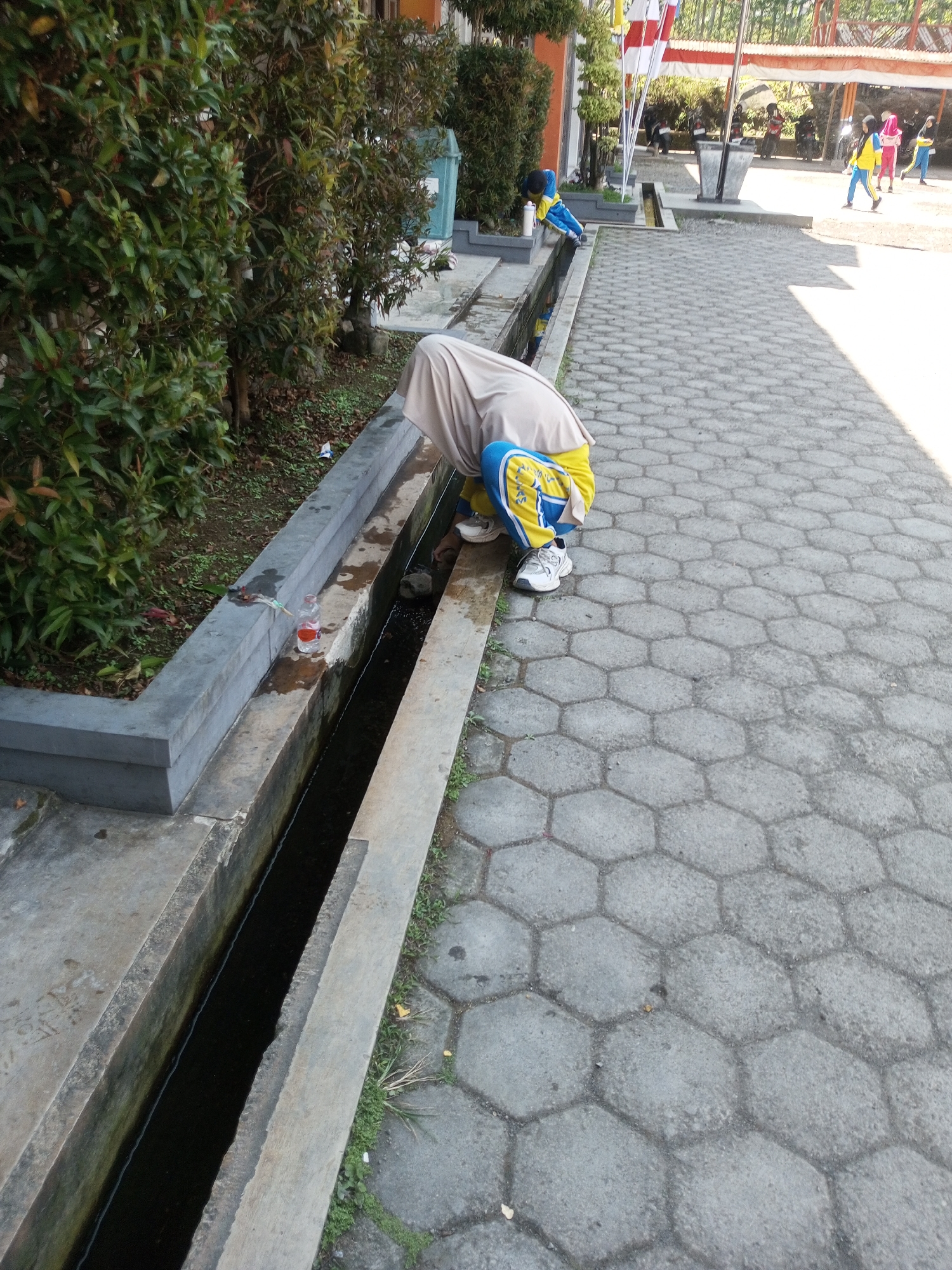 In the city of Kalibeber, Indonesia, an unidentified person wearing a light brown hijab and a yellow and blue school uniform crouches beside a concrete drainage ditch. The person is leaning over the ditch, with their right hand reaching into the dark water. A partially empty plastic bottle with a red lid, containing a small amount of liquid, rests on the edge of the ditch near the person. The ditch runs alongside a paved walkway made of hexagonal gray concrete tiles. Lush green bushes border the walkway and the ditch. In the background, other individuals in school uniforms are visible near a building. The scene appears to be an outdoor school or public area on a sunny day.