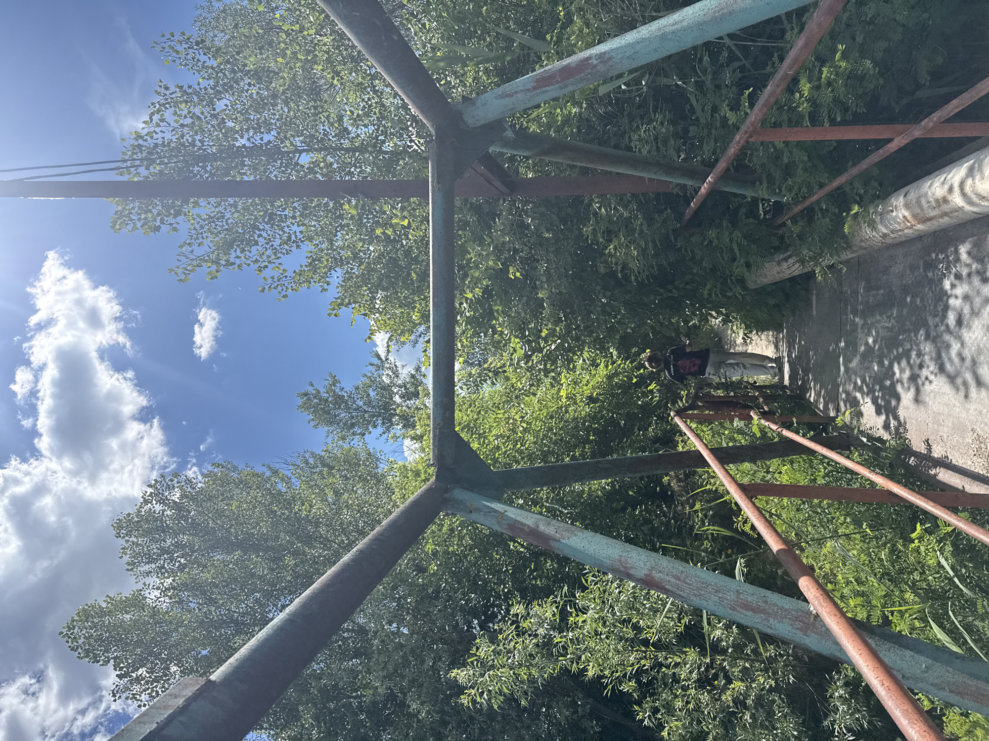 A person walks along an old, rusty metal bridge covered in overgrowth in Obukhivka, Ukraine. The bridge's structural beams are painted in faded teal and rust colors, with orange railings. Lush green trees and foliage surround the bridge, with a bright blue sky and scattered white clouds visible above. The ground of the bridge appears to be made of concrete or a similar material, with sunlight casting shadows. The person is seen from behind, wearing a dark t-shirt and light-colored pants, walking away from the camera towards the far end of the bridge.