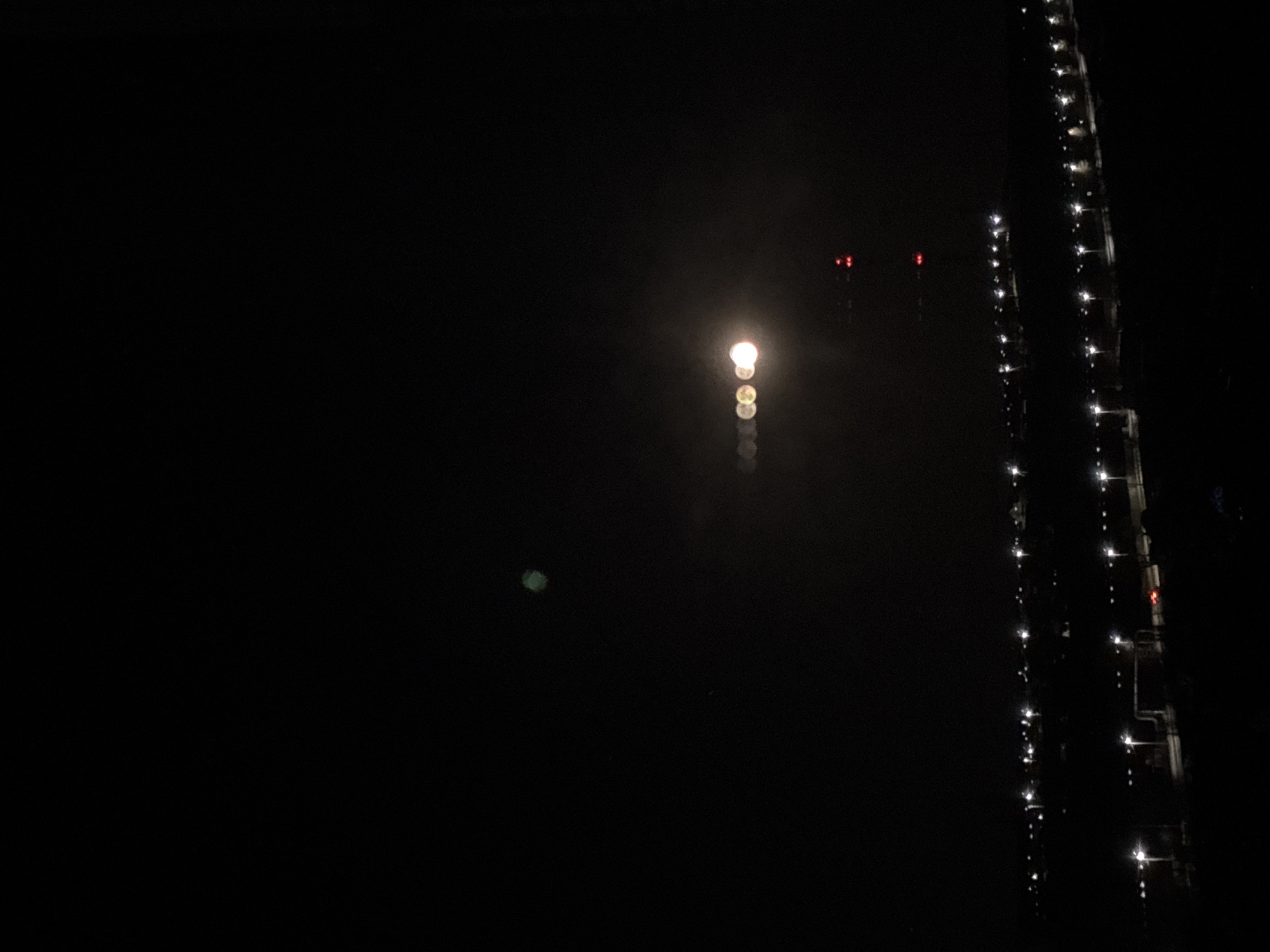 The image captures a night scene in Chaikovsky, Russia. A long structure, likely a pier or bridge, extends vertically on the right side of the frame, illuminated by evenly spaced lights. Further away, a single, bright light source is visible in the center, with blurred reflections in the water below. Two red lights appear in the background, potentially indicating structures or navigational aids. A faint green light is visible to the left of the main light source. The rest of the scene is predominantly dark, suggesting an absence of other light sources in the vicinity. The water surface appears calm and reflective.
