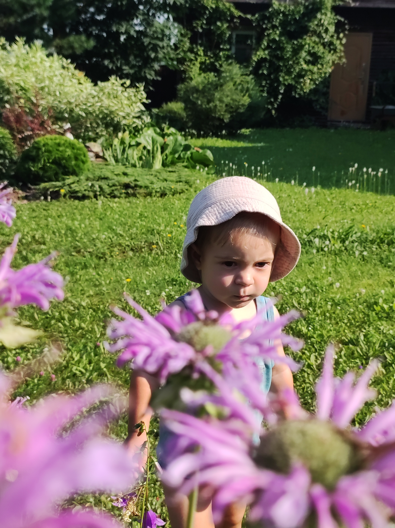 A young child, wearing a light-colored sun hat and blue overalls, stands in a grassy yard. The child is looking to the left of the frame with a neutral expression. In the foreground, out-of-focus purple flowers are prominent. Behind the child, the yard extends to a garden with various plants and shrubs, and a wooden building with a dark roof is visible in the background. The scene appears to be in the city of Fokino, Russia, based on the context provided.
