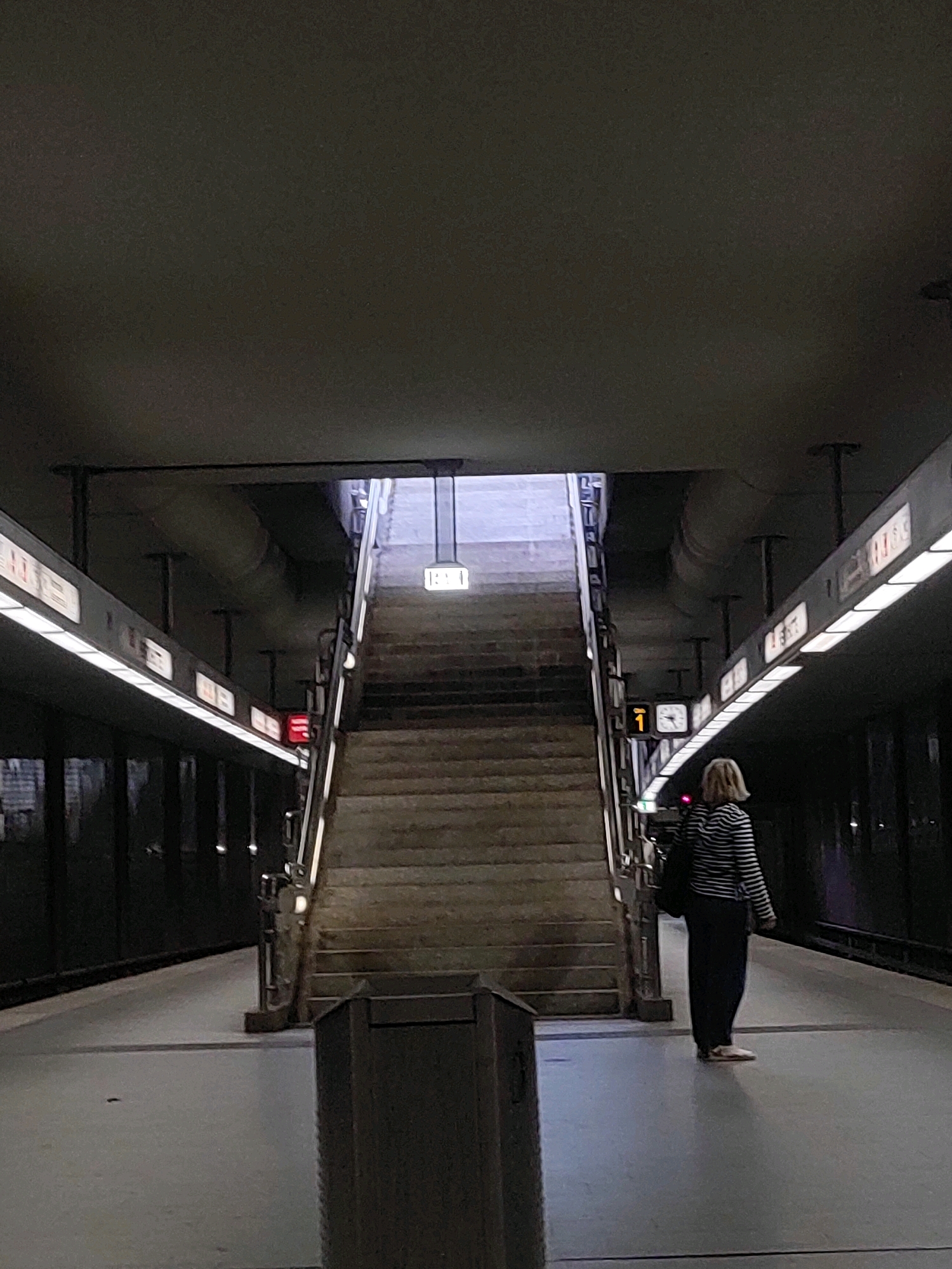 The image depicts a woman standing on a subway platform in Nuremberg, Germany. She is facing away from the camera, looking towards a long staircase that leads upwards and out of view. The woman is wearing a black and white striped long-sleeved shirt and dark pants. She has a backpack slung over her shoulder. To her right, a clock displays the time as approximately 1:05. Above the clock, a sign with the number "1" is visible, likely indicating a platform number. The platform is brightly lit by overhead lights and illuminated signs along the wall. The staircase is made of concrete and has metal railings. In the foreground, a dark trash receptacle is present. The overall environment is a modern subway station, characterized by concrete structures, metal fixtures, and artificial lighting.