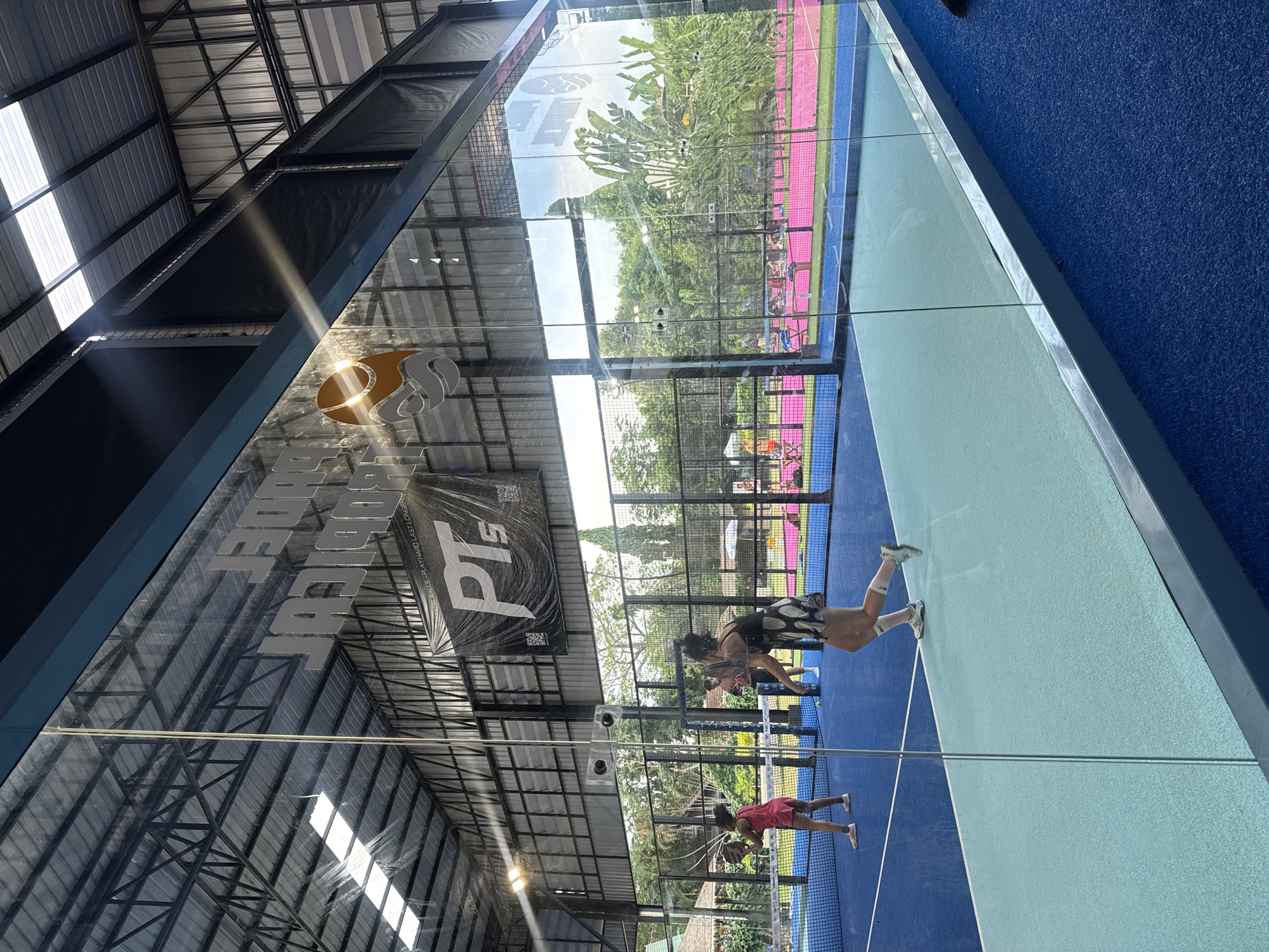 An indoor padel tennis court in Baan Bang Rak, Thailand. Two women are playing padel tennis on a blue court with a turquoise border. One woman in a pink outfit is on the left side of the net, holding a racket. The other woman in a black and white polka dot skirt and black top is on the right side of the net, mid-stride. A man is visible in the background near the net. The court is enclosed by glass walls and a metal frame. Above the court, a metal roof with skylights allows natural light to enter. A black banner with white lettering and a circular logo hangs from the ceiling. Trees and other vegetation are visible through the glass walls, indicating an outdoor environment surrounding the indoor facility.