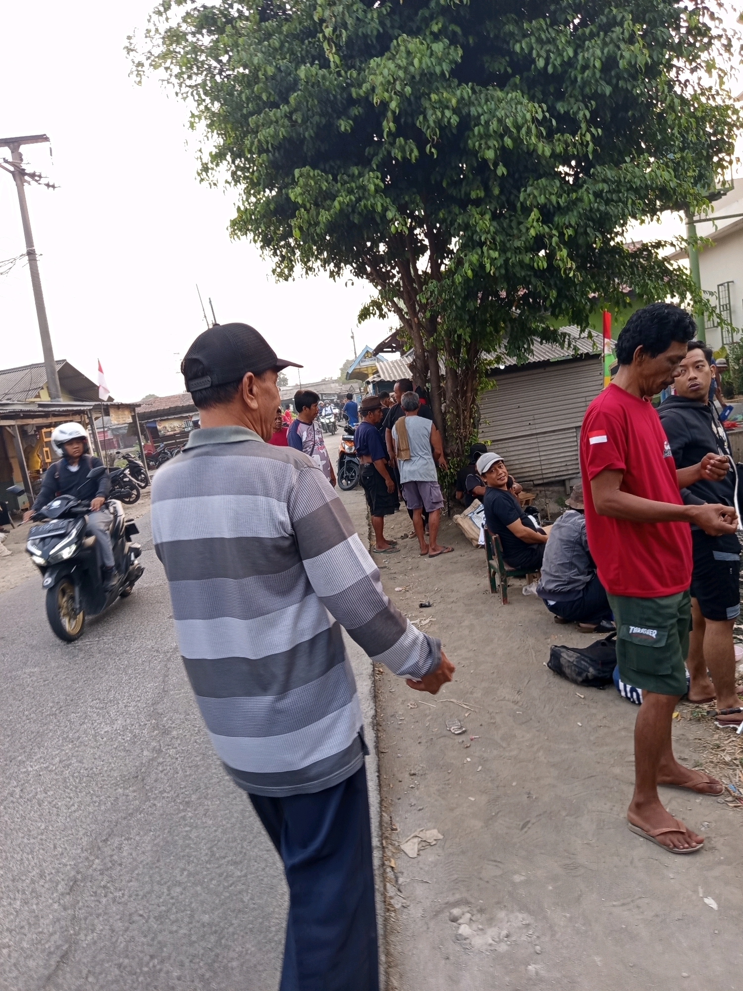 A group of men are gathered on a roadside in Karawang, Indonesia. In the foreground, a man wearing a grey and black horizontally striped long-sleeved shirt and dark blue pants turns away from the camera. To his right, a man in a red t-shirt with an Indonesian flag emblem and green shorts stands with his hands clasped. Behind him, another man wears a black hoodie and dark shorts. Several other men are visible in the background, some sitting on a low wall, others standing. A person on a black motorcycle wearing a white helmet is seen from the side, riding down the asphalt road on the left. The environment consists of a paved road, a dirt shoulder, and a line of buildings with corrugated metal roofs. A large tree with dense green foliage is situated behind the gathering. A utility pole and wires are present on the left side of the frame. The sky is overcast.
