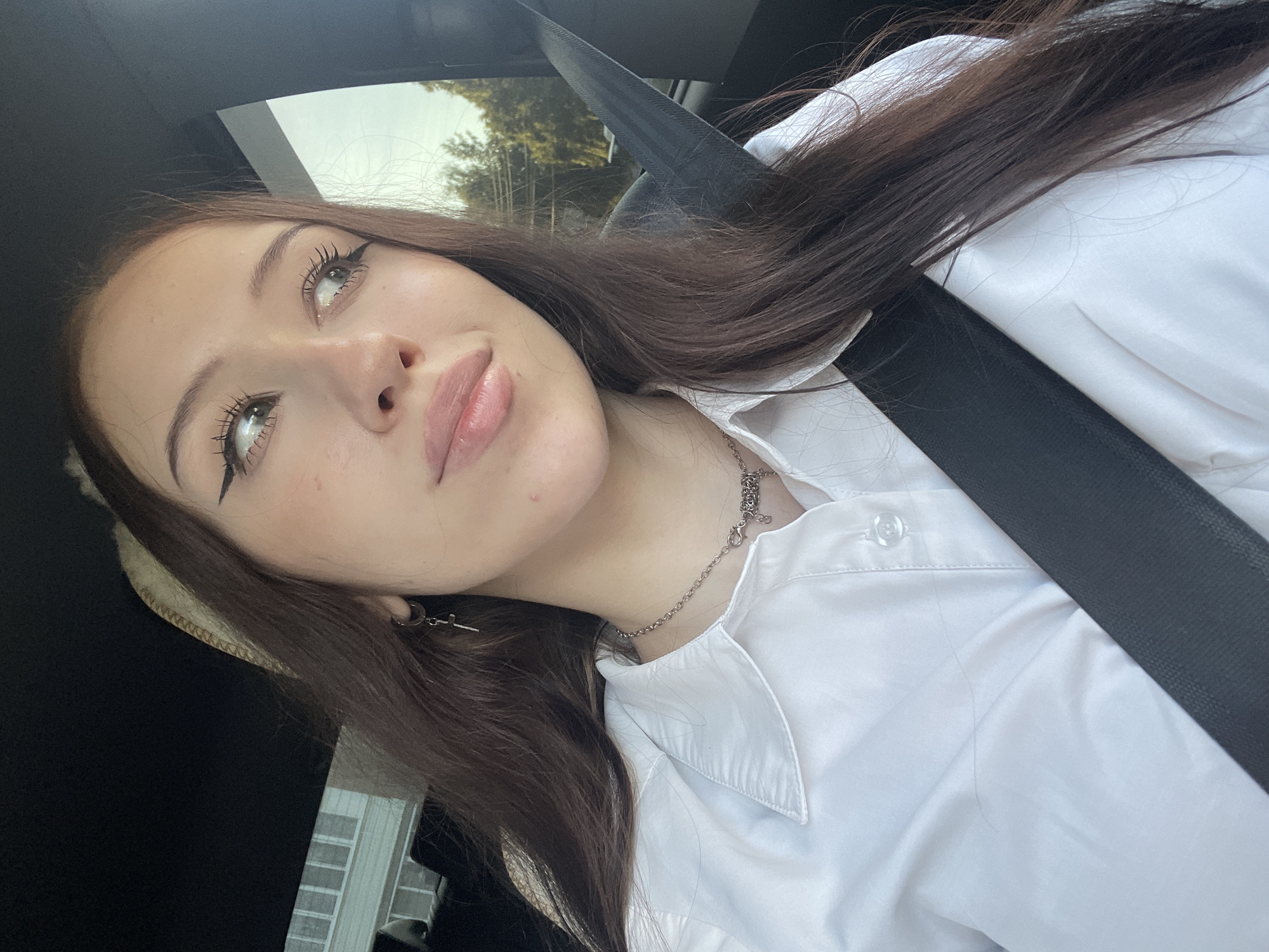 A young woman with long, dark brown hair and light skin is pictured from the chest up, seated in a car. She is wearing a white button-down shirt and a silver necklace with a charm. A black seatbelt crosses her chest. Her eye makeup is prominent, featuring winged eyeliner and long lashes. Her gaze is directed away from the camera, upwards and to her right. The background is blurred, showing a residential building with light-colored siding on the left and trees and a bright sky on the right. The interior of the car, including a dark headliner, is visible at the top of the frame. There is no specific indication of the location within the city of Votkinsk, Russia.