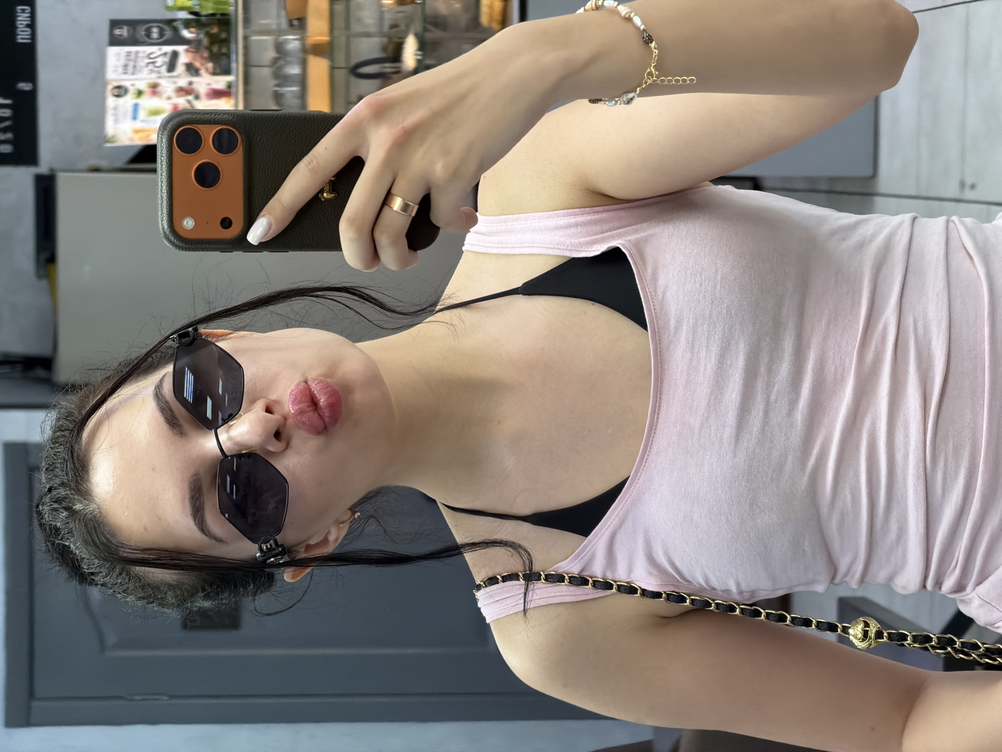 A young woman with dark hair pulled back is taking a selfie in what appears to be a retail environment. She is wearing sunglasses, a pink tank top over a black bra, a gold ring on her left ring finger, and a bracelet on her right wrist. She is holding a smartphone in her right hand, which has an orange and black case. Her lips are pursed as if blowing a kiss. The background shows shelves with various items and a doorway.