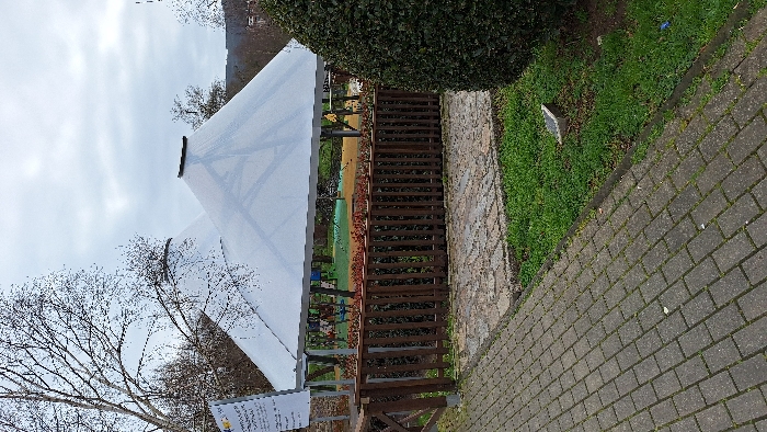 An outdoor scene in Arteixo, Spain, presents a section of a playground facility. In the immediate foreground, a grey paver pathway extends diagonally from the lower left, bordered on its right by a strip of green grass containing small debris. A large, dark green, manicured evergreen bush occupies the lower right quadrant. A dark brown wooden fence with vertical slats and a horizontal top rail, adorned with small reddish-brown plants, separates the foreground from the main area. Beyond this fence, two prominent, light-colored, translucent conical canopy structures, supported by metal frameworks, cover a section of a playground. Under these canopies, various colorful playground equipment, including blue and yellow swings, are visible against a reddish-orange soft-fall ground. To the left of the scene, a white rectangular sign mounted on a pole displays unreadable text. Bare deciduous trees with intricate branch patterns are visible in the background and between the canopies, silhouetted against an overcast, light grey sky. No individuals are present in the scene.