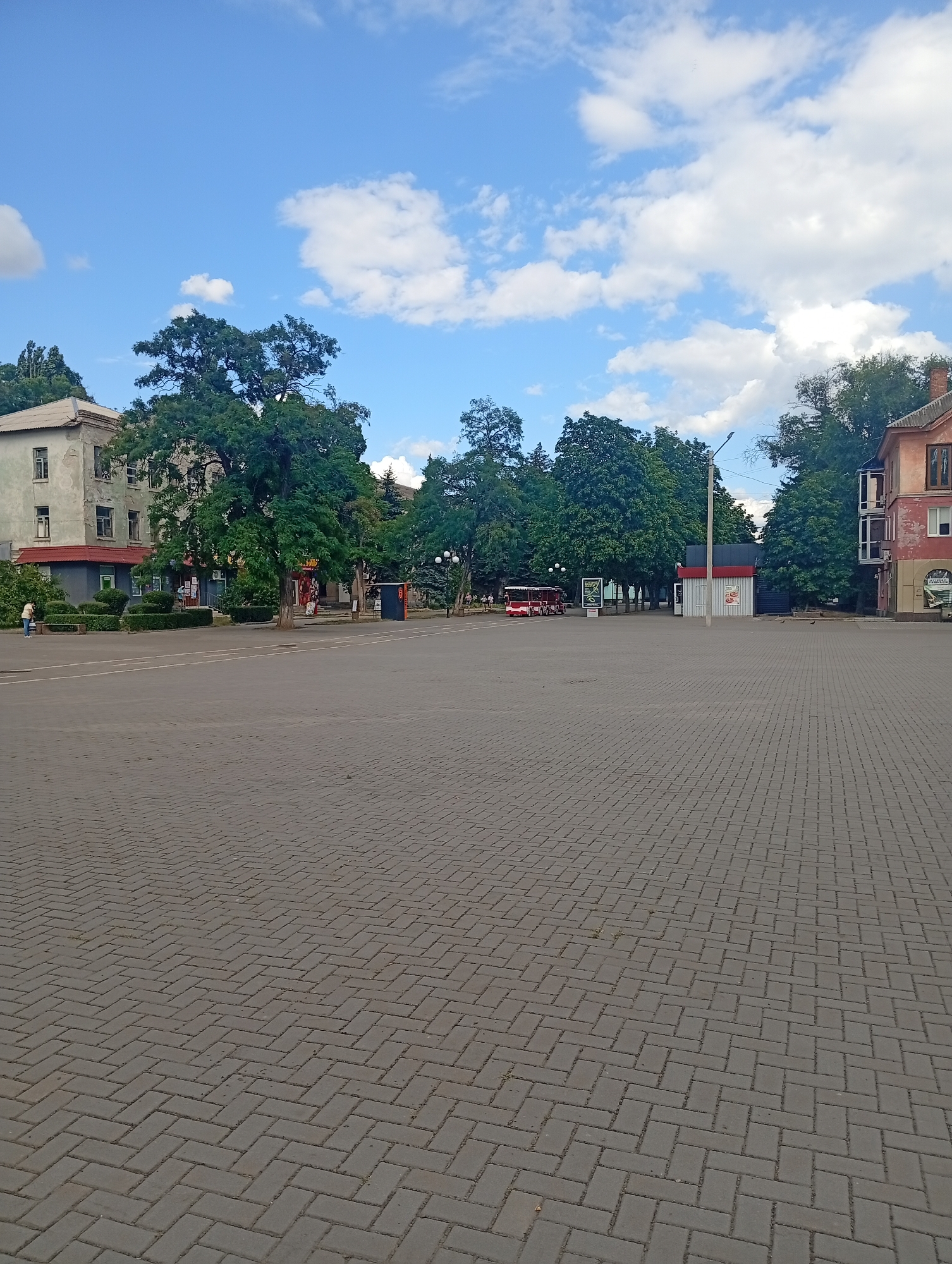This is an outdoor, eye-level, full-shot photograph of a paved town square in Pokrov, Ukraine. The square is made of grey interlocking paving stones laid in a herringbone pattern. The foreground is dominated by the paving stones, which recede into the distance. In the middle ground, there is a red tour bus parked on the left side of the square. Behind the bus, there are several buildings and trees. On the left side of the square, there is a grey, multi-story building with windows. In front of it, there are some low, green bushes and a bench. A person in a light-colored jacket is standing near the bench, facing away from the camera. Further back, there is a small red and white kiosk. On the right side of the square, there is a multi-story building with a reddish-brown facade and windows. There are trees scattered throughout the middle and background of the image, with lush green foliage. The sky is blue with scattered white clouds. The scene appears to be a public space, possibly a city center or a plaza.