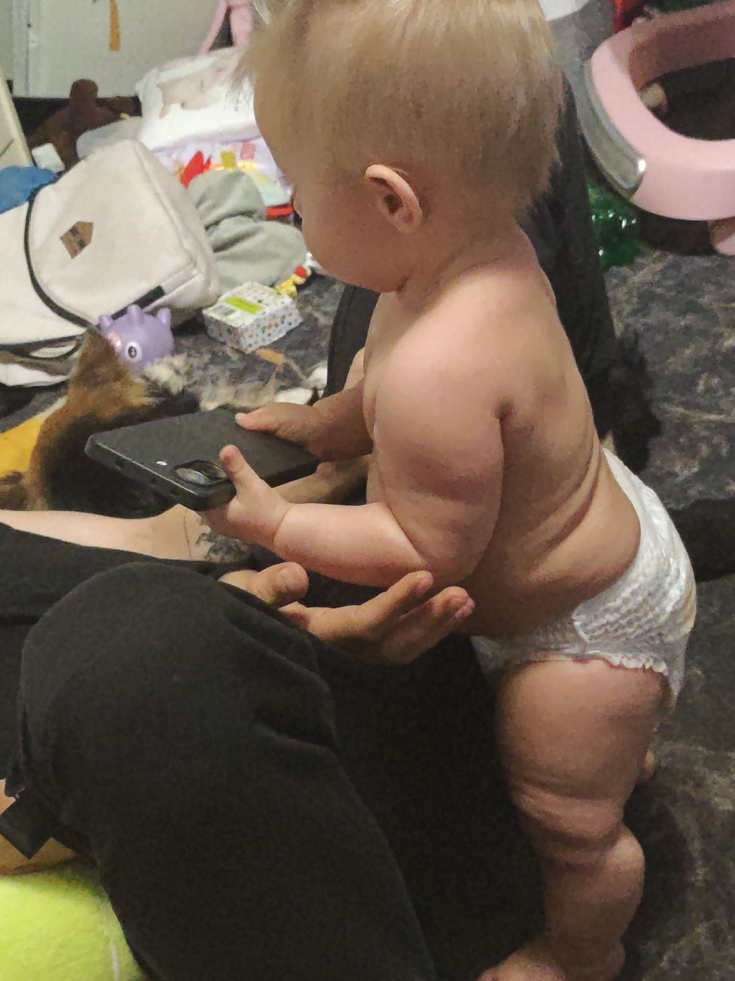 The image shows a toddler standing and holding a black smartphone. The toddler is wearing a diaper. An adult's hands are visible, one holding the phone, the other supporting the toddler. The background appears to be a cluttered indoor environment with various objects such as bags, toys, and what might be bedding. The location is not specified in the image or provided crops.