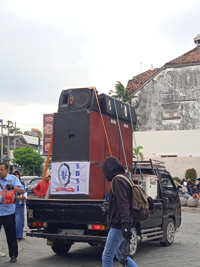 The image shows a black pickup truck parked on a paved street in Yogyakarta, Indonesia (latitude: -7.783, longitude: 110.367). The truck is carrying a stack of large, rectangular speakers secured with orange straps. One person is wearing a black hoodie and jeans and holding a black object that could be a tripod or a long, black, cylindrical object. A white banner is displayed on the truck's side with the letters "SBSI" and a logo featuring the Indonesian flag. Another person is wearing a red shirt, and a third is wearing a blue shirt with a red vest and carrying a red bag. The background shows a grey building with a window and a smaller, red-roofed building in front of it. A sign for "Mandiri" and another for "ASIN" are visible behind the truck. Other pedestrians are out of focus. The scene takes place on a street with minimal traffic and is overcast.