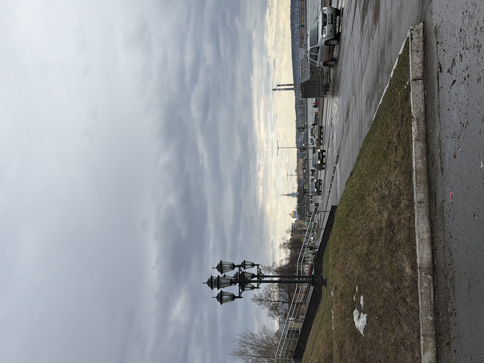 The image displays an overcast daytime scene in an urban or semi-urban setting, identified as Votkinsk, Russia. The perspective is from a street level, looking across a road towards a distant landscape.
In the immediate foreground, a wet asphalt road occupies the right side of the frame, indicating recent rain or melting snow. It is bordered by a rough concrete curb, beyond which lies a sloping grassy bank with visible patches of melting snow, suggesting a cool season like late winter or early spring. A prominent feature on this bank is a tall, decorative black street lamp with six ornate lanterns. A metal railing runs along the bank's edge, separating it from a lower elevation or another section of the road.
On the main road, several vehicles are moving, denoting ongoing traffic. A silver SUV, possibly a Ford, is clearly visible, with a partially discernible Russian license plate: H711XX 152. The region code '152' typically belongs to Nizhny Novgorod Oblast. Further down the road, other cars and what appears to be a truck or trailer can be seen, along with green traffic lights.
The background showcases a distant landscape, potentially across a body of water or a wide valley, where a town or city is visible. A distinctive white building topped with a prominent golden dome, resembling a church, stands out. To its left, two tall, slender industrial chimneys pierce the horizon. The sky is entirely covered by thick, uniform grey clouds, creating a subdued and somber atmosphere. No individuals are clearly visible, and no specific event is taking place beyond the routine movement of traffic.