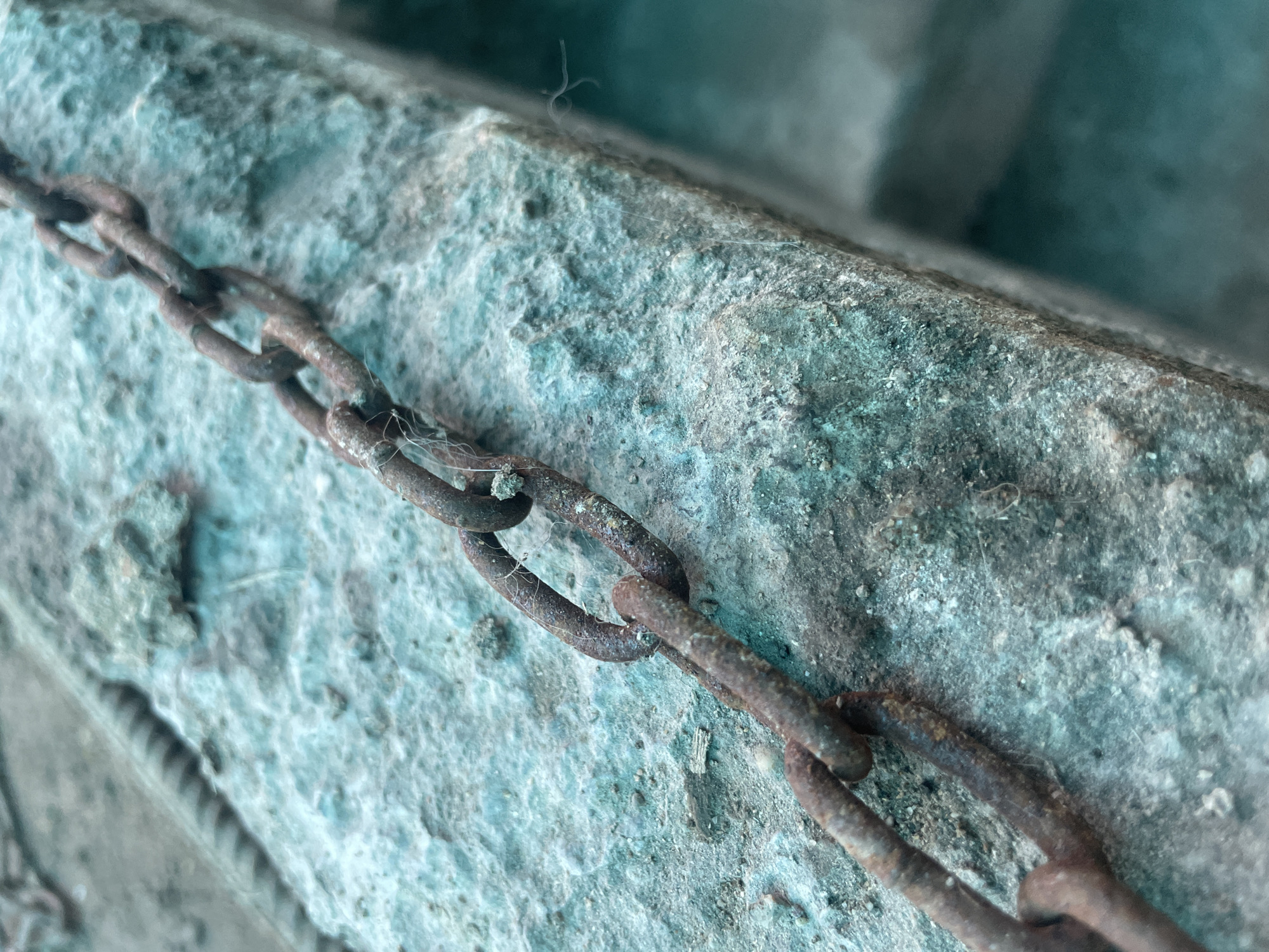 The image shows a close-up of a rusty metal chain lying on a rough, textured surface, possibly concrete or stone. The surface has a blue-green hue. The chain shows signs of corrosion. The scene appears to be outdoors. The location is Olginka, Russia.
