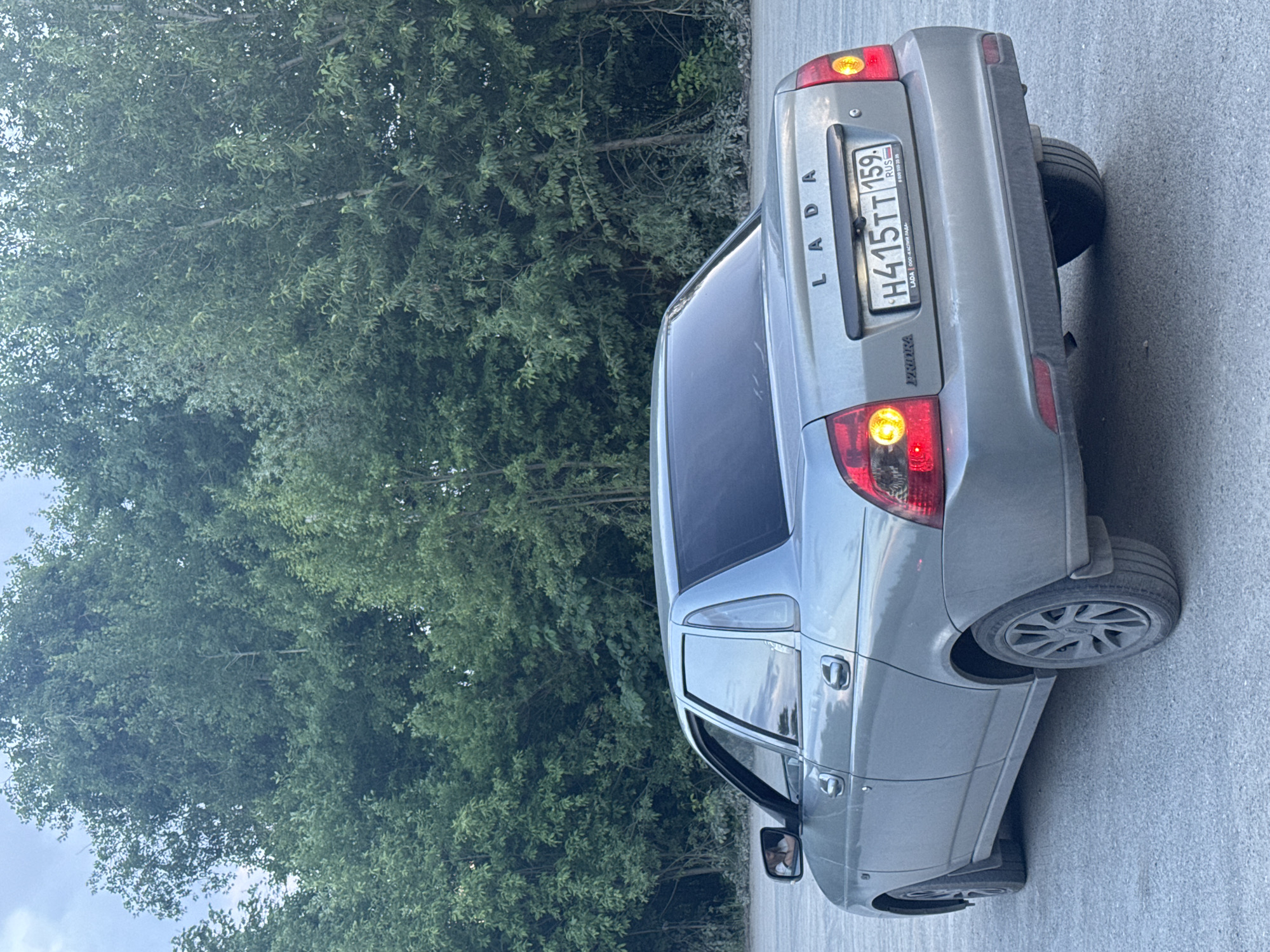 A light grey Lada Priora sedan is parked on a paved surface, likely a parking lot, with a dense line of green trees and foliage forming the background. The car's rear is angled towards the viewer, showcasing its taillights, bumper, and license plate. The license plate reads "H415TT 159 RUS". The car appears to be dusty. In the driver's side rearview mirror, a partial reflection of a person is visible. The sky above is overcast with a muted light. The overall environment suggests an outdoor setting, possibly on the outskirts of a city or in a park area.