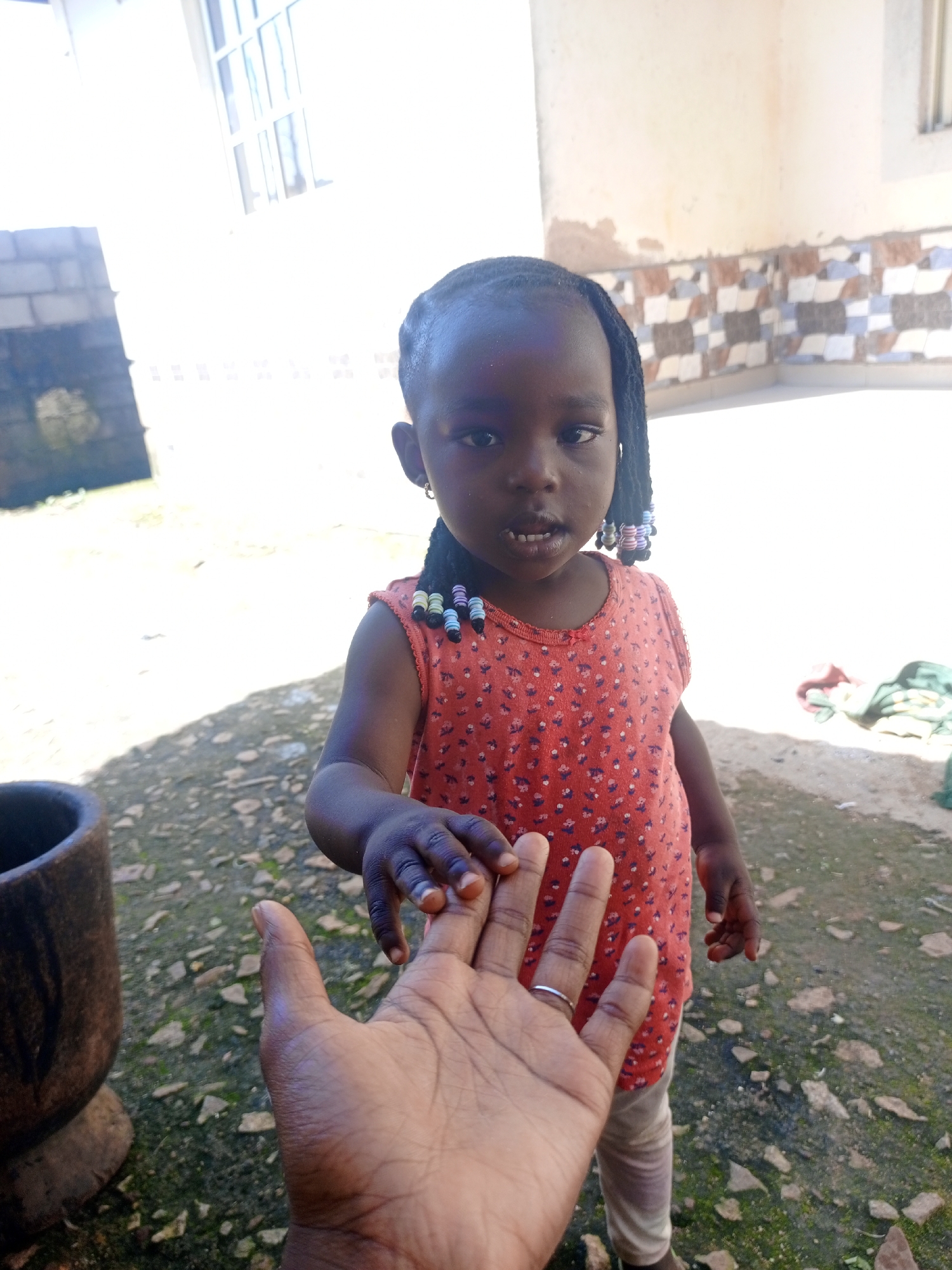 A young Black girl with dark skin and braided hair, adorned with colorful beads, stands outdoors. She wears a coral-colored sleeveless shirt with a floral pattern and light-colored pants. Her left hand is outstretched and grasps the finger of an adult's hand, which is in the foreground. The adult's hand wears a silver ring on the ring finger. Behind the girl, a white building with windows and a tiled lower section is visible. To the left, a weathered stone mortar is present. The ground is covered with stones and some patches of green. The lighting suggests it is daytime, with bright sunlight casting shadows. The scene appears to be in a non-urban setting or a less developed area of a city.