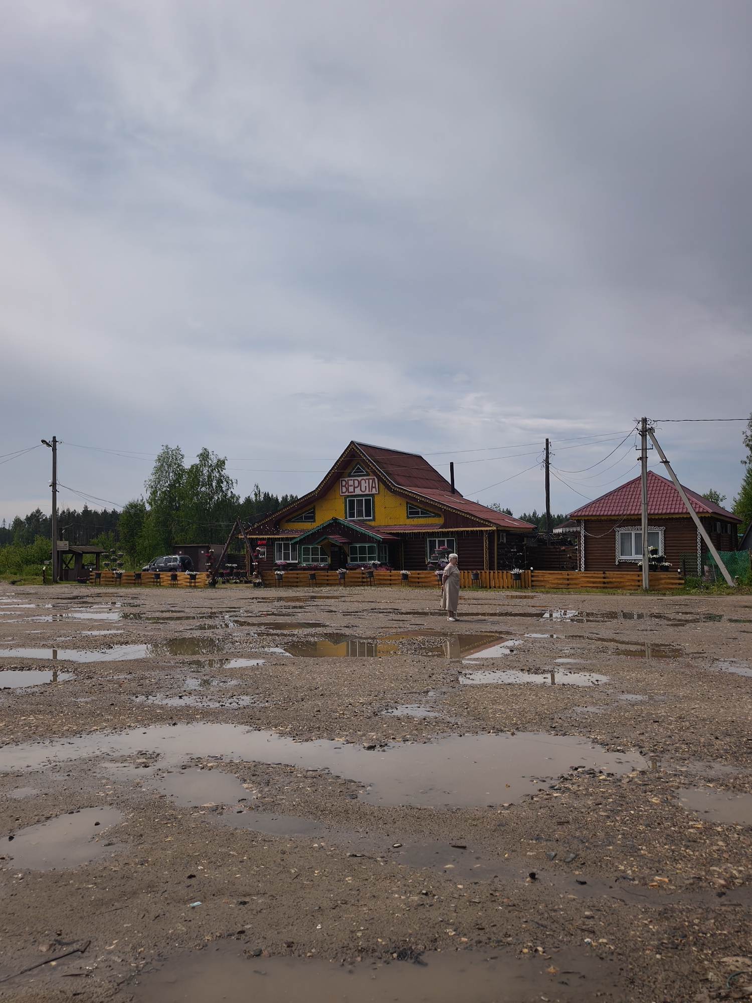 The image depicts an outdoor scene on a cloudy day. The foreground is dominated by a muddy, gravelly ground with numerous large puddles, indicating recent rainfall or poor drainage. In the mid-ground, a cluster of wooden buildings is visible. The largest building, painted yellow with a red roof, has "ВЕРСТА" written in red on its facade. A smaller, darker wooden building with a red roof stands to its right. A single person, dressed in a long, light-colored robe and head covering, stands between the buildings, facing away from the camera. The background features a line of trees and a forest. Electrical poles and wires are present in the scene, suggesting the presence of infrastructure. The overall impression is one of a rural or semi-rural location, possibly a roadside establishment or a small village. Based on the Cyrillic signage, the location is in Russia. Specifically, this scene appears to be at the roadside establishment "Верста" (Versta) located in the village of Kady, Kologrivsky District, Kostroma Oblast, Russia.