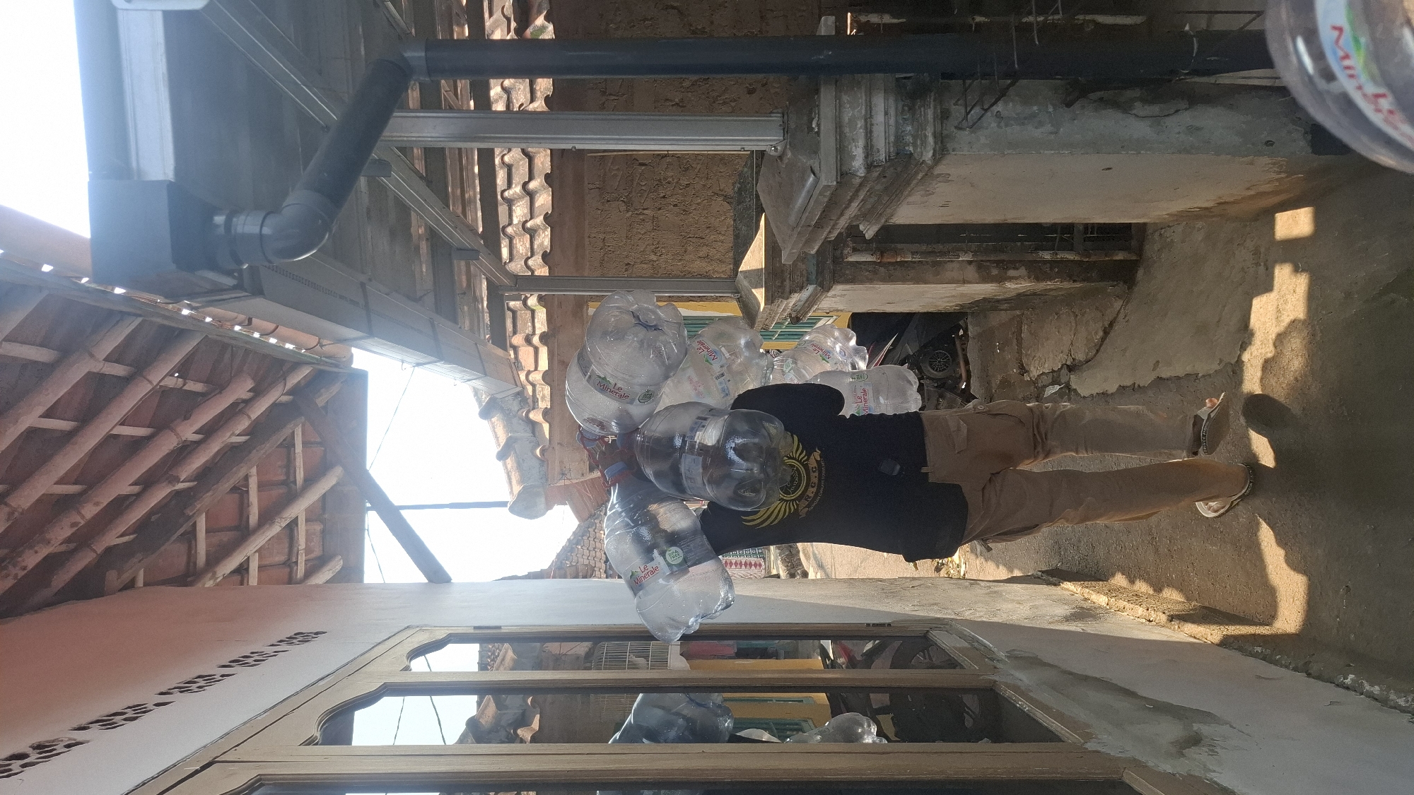 A person is walking down a narrow alleyway in Rahong, Indonesia, carrying a large load of bottled water on their shoulders. The person is wearing a black t-shirt and khaki pants, and flip-flops. The alleyway is made of concrete and is lined with buildings. The buildings have red tile roofs and clay walls. The sun is shining brightly, casting shadows on the ground. There is a gutter and downspout on the right side of the alley. The person is carrying multiple large plastic bottles of water, some of which appear to be branded "Le Minerale". The bottles are gathered together with some kind of strap or handle. The person's back is to the camera.