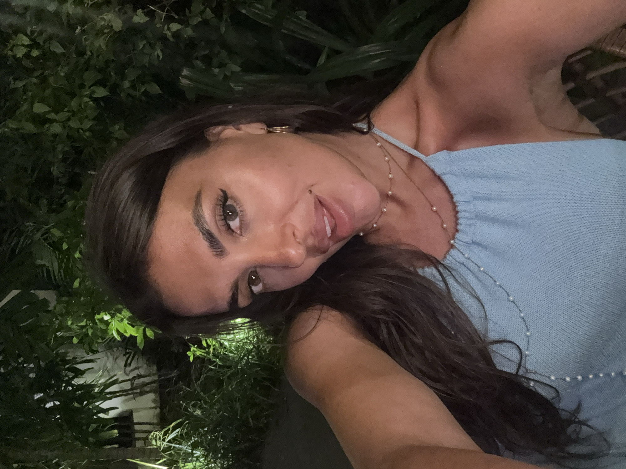 A medium shot depicts a woman with dark hair, light skin, and prominent eyebrows, wearing a light blue top with pearl embellishments. She is positioned against a backdrop of dense green foliage, possibly indicative of tropical vegetation. The environment is dimly lit, suggesting an evening setting. The location is inferred to be Kuta Selatan, Indonesia, based on the prompt.