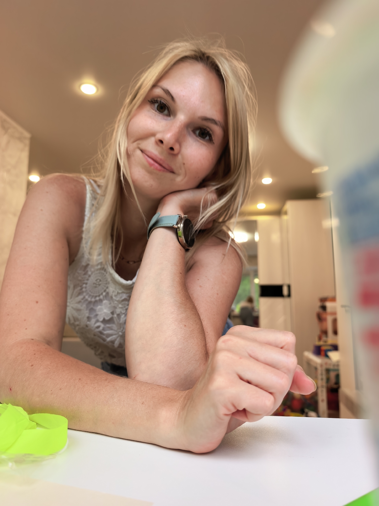 A young Caucasian woman with blonde hair and fair skin is captured in a close-up, waist-up shot. She is leaning on her arm, with her chin resting on her hand, and she is looking directly at the camera with a slight smile. She is wearing a white, lacy tank top and a watch on her left wrist. The background appears to be an indoor setting, possibly a kitchen, with white cabinets and lighting visible. A blurred object, possibly a large container, is in the foreground on the right side of the image. A bright neon green object, likely a rubber band or similar item, is on the left side of the image. The woman's skin shows some freckles. There is no visible indication of the city of Sterlitamak, Russia.