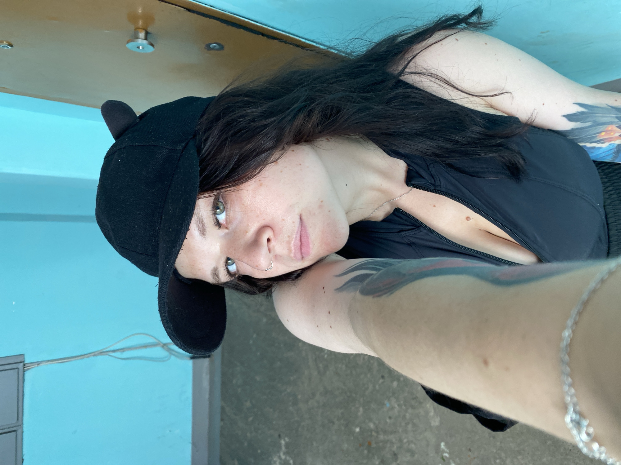A young woman with dark hair, freckles, and green eyes is sitting on the floor, leaning against a blue wall. She is wearing a black baseball cap with cat ears, a black zip-up top, and black shorts. She has tattoos on her arms and a silver bracelet on her wrist. The room appears to be an indoor space with a concrete floor and a wooden door. The lighting is bright, suggesting it may be daytime. The setting is consistent with an apartment building or similar residential structure in Chaikovsky, Russia.
