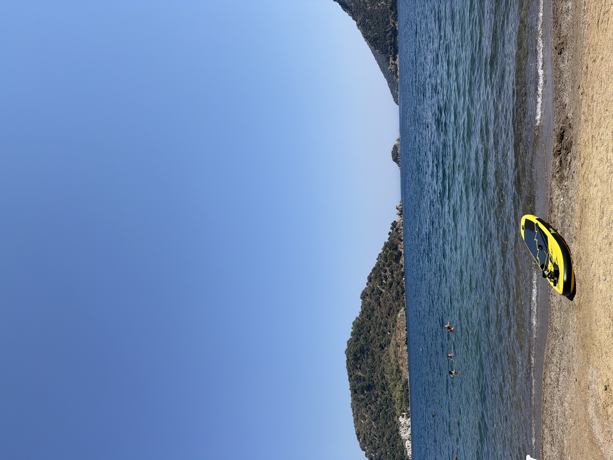 A bright, sunny day showcases a scenic view of Marmaris, Turkey's coastline. The foreground is a sandy beach with a bright yellow and black stand-up paddleboard resting on the sand near the water's edge. The clear blue water laps gently onto the shore. In the middle ground, several people are swimming or wading in the sea. The background features lush, green, tree-covered hills that form the coastline, with a few small islands visible in the distance. The sky is a clear, vibrant blue.