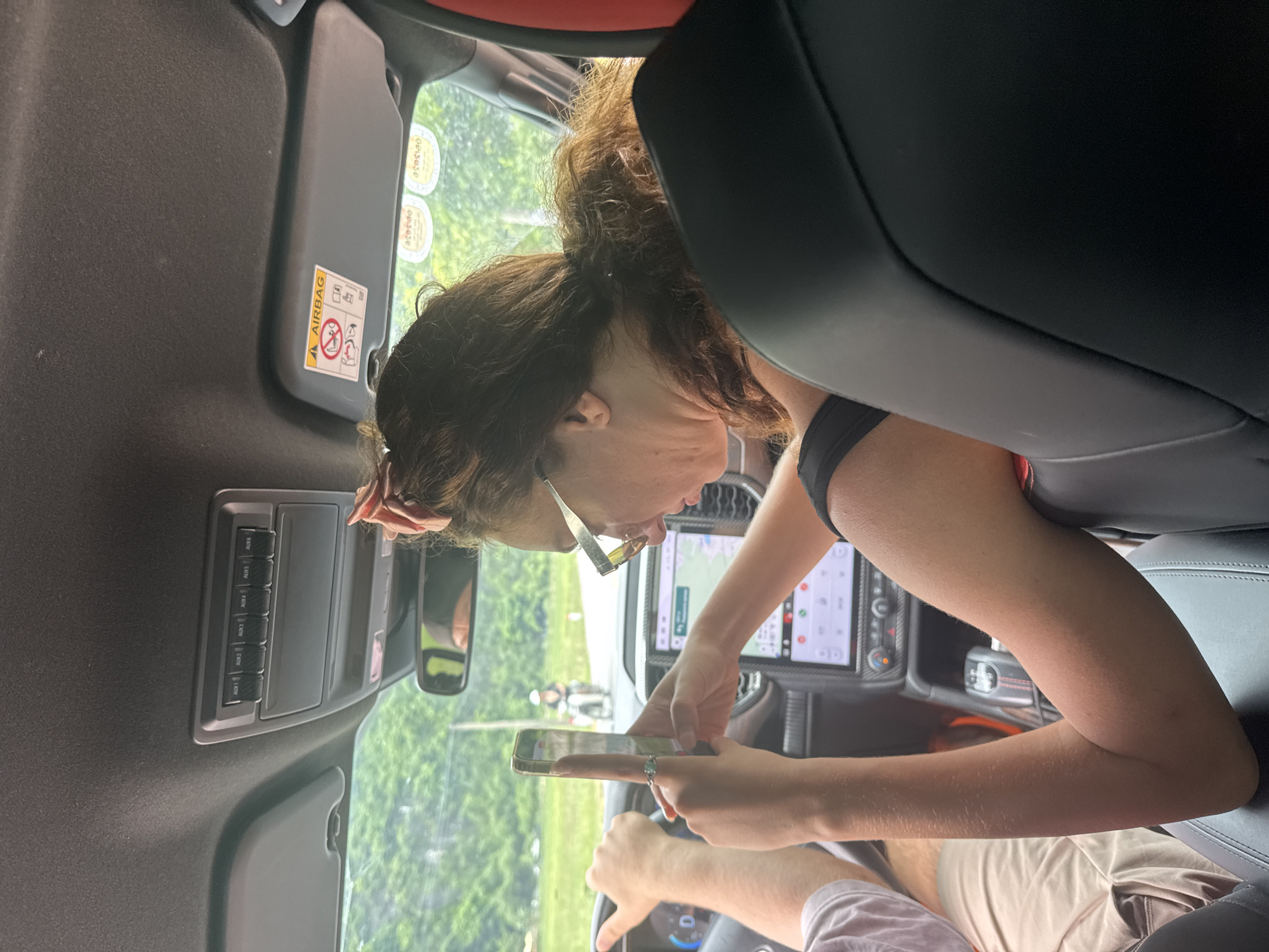 The image depicts the interior of a vehicle, likely in the city of Nam Hoa Lư Ward, Vietnam. Two individuals are visible. In the foreground, a woman with long, curly brown hair tied back with a pink bow is wearing glasses and looking at her phone. She is in the passenger seat. Her left arm is resting on the seat. In the driver's seat, a person's hands are on the steering wheel. The car's dashboard displays a navigation screen. Outside the vehicle, a road is visible with greenery and trees lining the sides. A motorcycle with a rider is in the distance on the road. The car's overhead console has buttons labeled "AUX 1" through "AUX 6," and a sun visor with an airbag warning sticker is visible.