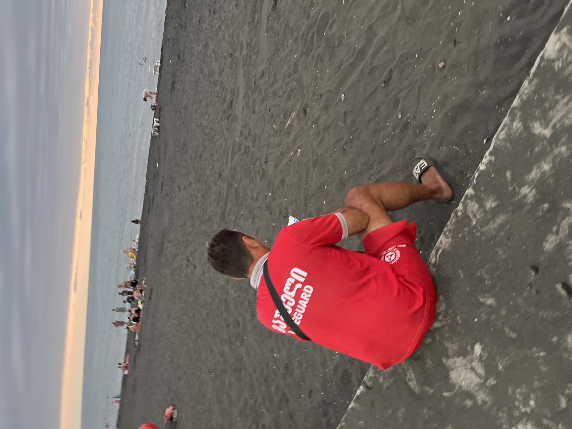 The image captures a scene on the black sand beach of Ureki, Georgia, with the Black Sea in the background. The sky is overcast with a hint of sunset color on the horizon. A man, identified by the text on his red shirt as a lifeguard, sits on a concrete embankment facing the water and looking at his phone. His uniform is red with "LIFEGUARD" written in white letters on the back. Other beachgoers are visible in the distance, scattered along the shoreline and in the water. Some are sitting or lying on the sand, while others appear to be wading. Beach furniture, such as lounge chairs, is also present. The overall atmosphere suggests a relaxed evening on the beach.