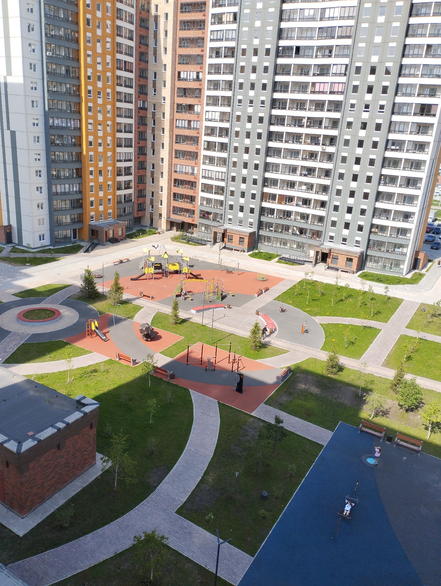 The image shows an aerial view of a courtyard in the city of Sadovyy, Russia. The courtyard is surrounded by tall apartment buildings with varying exterior colors and designs. A playground with colorful equipment and a red surface is situated in the center of the courtyard. There are multiple benches, small trees, and grassy areas throughout the courtyard. Walkways made of light-colored paving stones connect different parts of the courtyard. In the lower right corner, there is a dark blue area with exercise equipment and two people. In the lower left corner, there is a brick building with a dark roof.