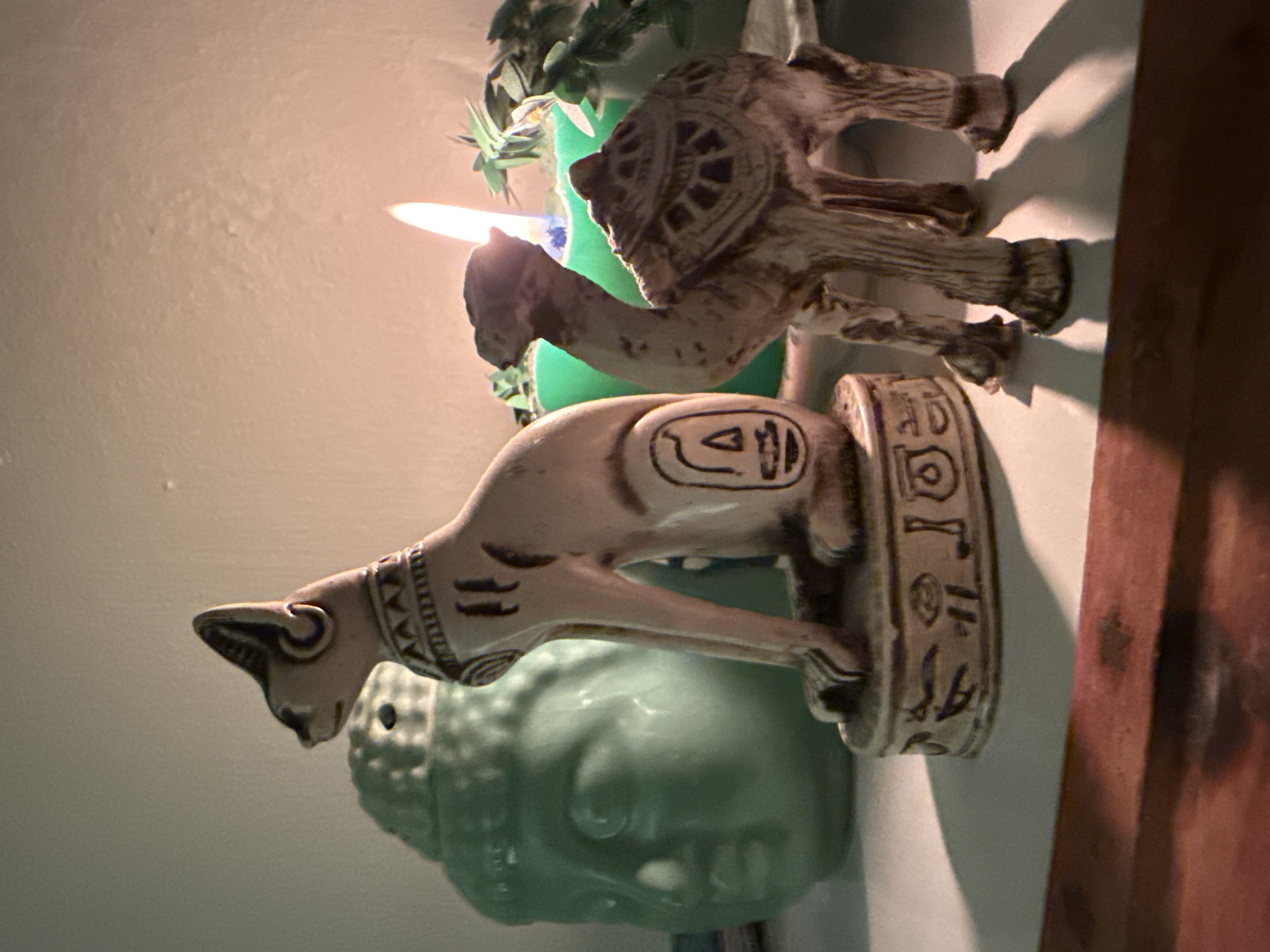 The image displays a still life composition featuring several decorative objects on a shelf. Dominating the foreground are two figurines: a stylized cat, likely representing the Egyptian deity Bastet, seated on a pedestal inscribed with hieroglyphs, and a standing camel. Behind these figures, a large, mint-green, Buddha-head-shaped ceramic object is visible to the left. To the right of the cat and camel, a lit green candle casts a warm glow, illuminating the scene and casting shadows. A few sprigs of greenery are placed behind the candle. The background consists of a textured, pale wall. The objects are arranged on a white surface, which appears to be a shelf, with a dark wooden surface visible below it. The lighting suggests an indoor setting, possibly at dusk or night, given the prominence of the candle flame. There are no people or animals present in the image. The scene is devoid of any specific location markers that would indicate Kadıköy, Turkey.