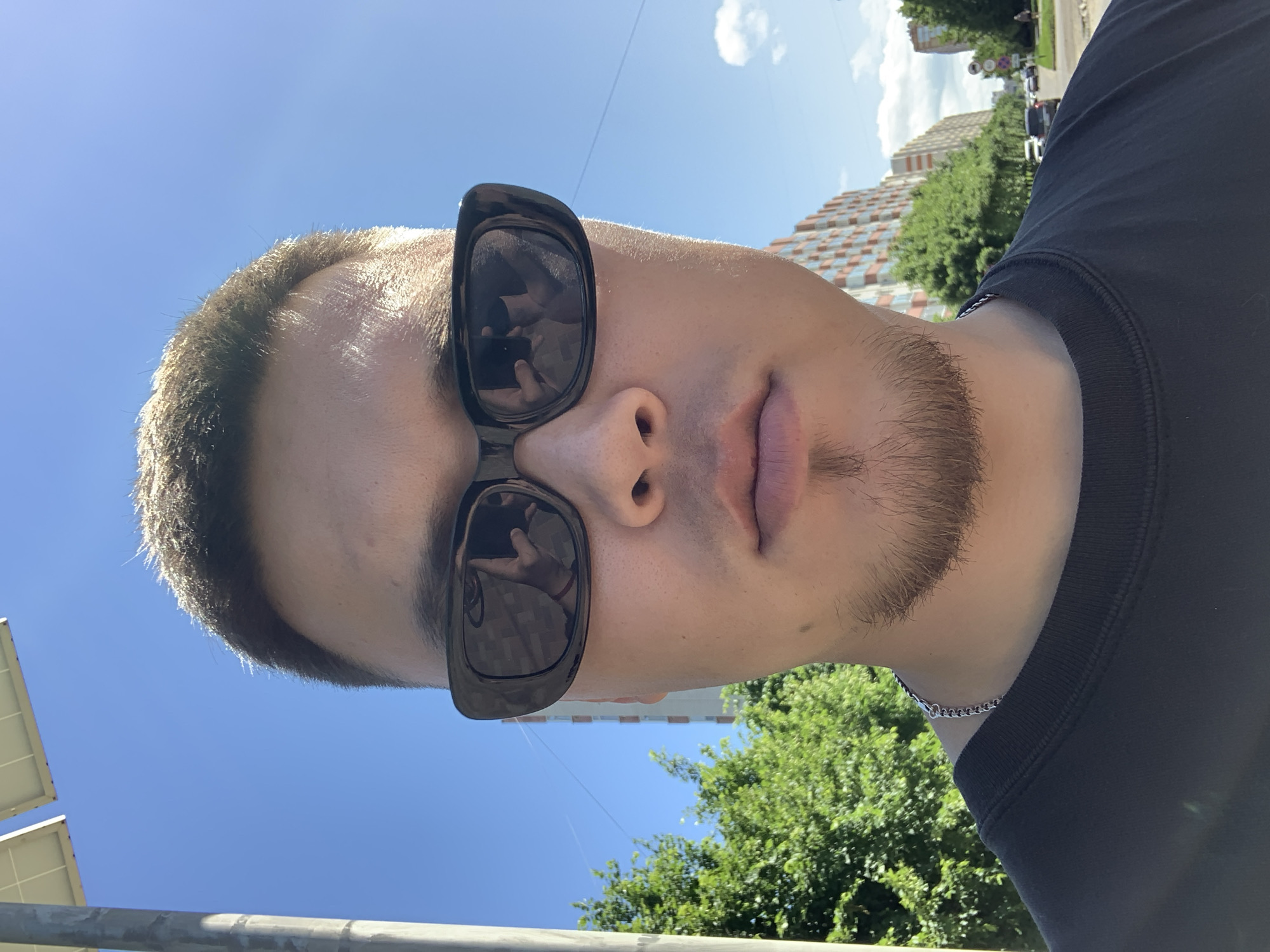 This is a close-up, upward-facing selfie of a young man outdoors. He is wearing large, dark sunglasses and a black t-shirt. His hair is short, and he has a light goatee. A silver chain is visible around his neck. The background shows a bright blue sky with a few scattered clouds, suggesting it is daytime and likely sunny. Green trees and a multi-story building with an orange facade are visible, indicating an urban or suburban setting. The building's architectural style and the presence of streetlights and other vehicles in the distance might suggest a location in Stavropol, Russia. The image appears to have been taken on a clear, warm day. The man's expression is neutral, with his lips slightly parted. The overall mood is casual and candid. There is no visible text in the image.