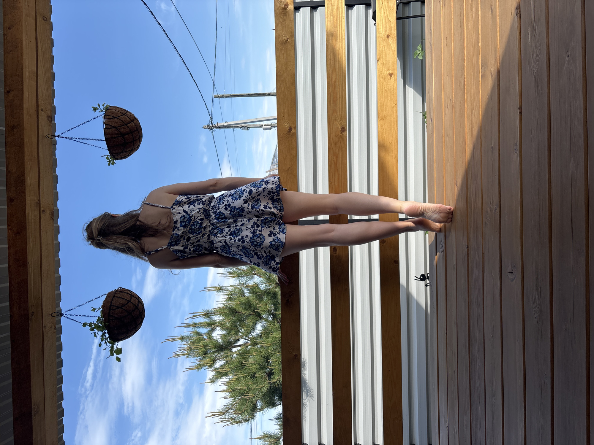 A woman stands on a wooden deck in Chaikovsky, Russia. She wears a blue and white floral dress. Two hanging baskets with greenery hang above her. A corrugated metal fence with wooden supports is visible behind her. A pine tree is in the background. Power lines cross the blue sky.