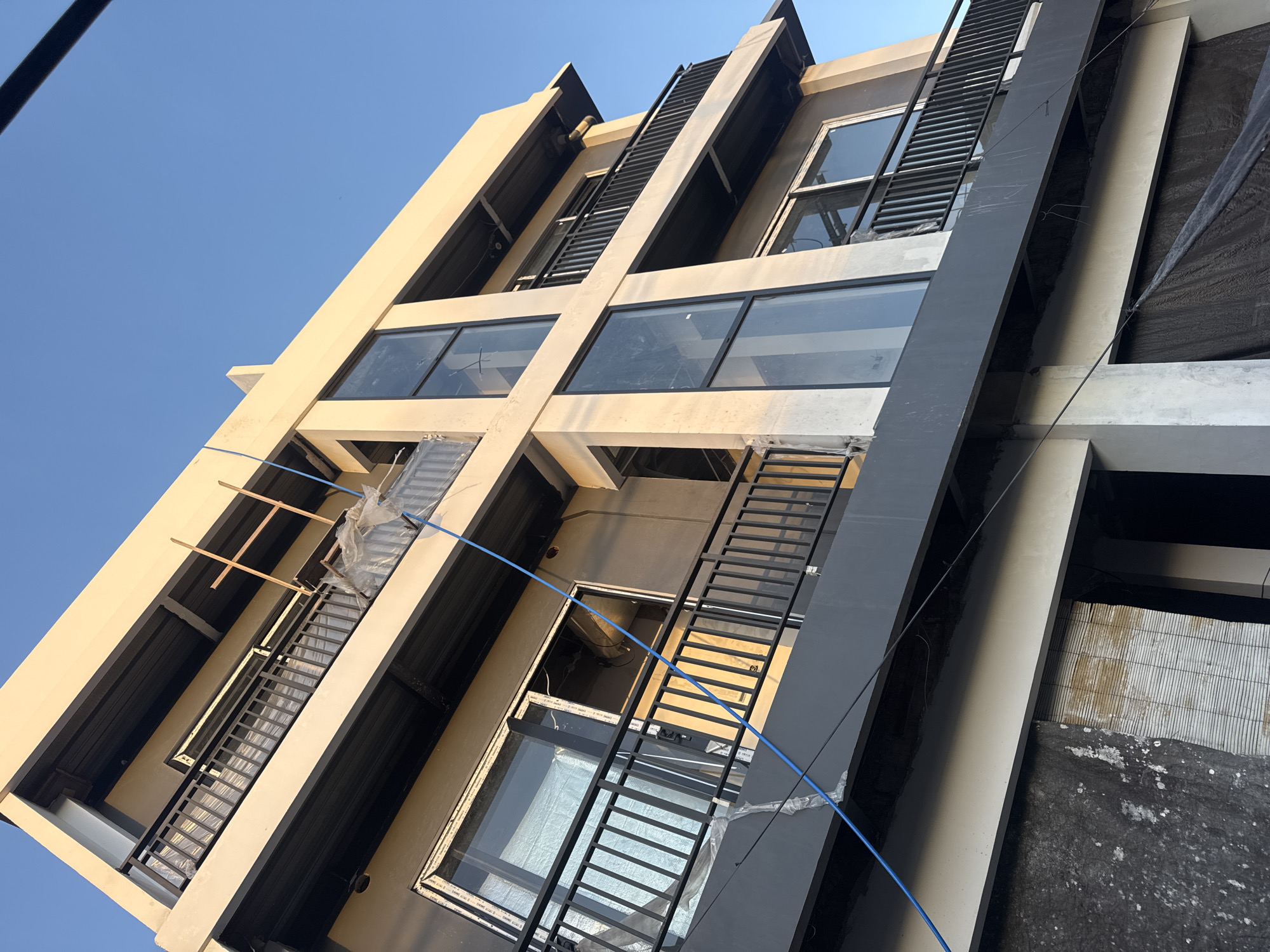 The image shows a multistory building under construction in Kuta Selatan, Indonesia, as deduced from the architectural style common in the region. The building features a modern design with a mix of light beige and dark gray vertical sections. Large windows are installed on each floor. Black metal ladders run vertically along the facade. Construction materials are visible within and around the windows, indicating ongoing work. A blue cable runs diagonally across the facade. The sky is clear and blue.