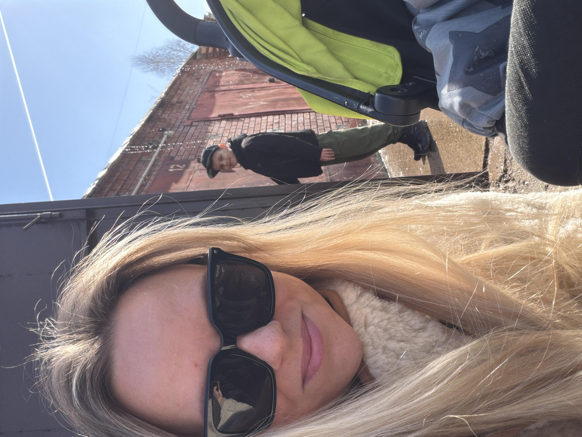 The photograph depicts a woman taking a selfie, with her face partially obscured by large sunglasses. She has long, blonde hair and is wearing a fluffy, light-colored coat or jacket. Behind her, reflected in a shiny surface, is a boy wearing a black jacket and cap, standing in front of a red brick building. There is also a stroller with a light green hood visible. The photograph appears to be taken outdoors on a sunny day. The location is identified as Votkinsk, Russia, based on the reflection of the building.