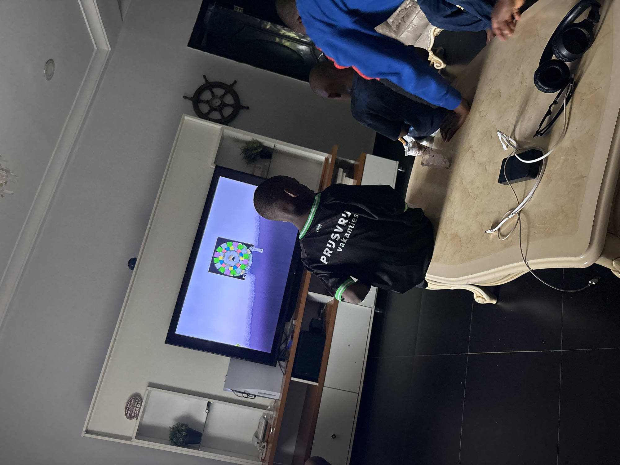 In a dimly lit room in Iddo Tudunwada, Nigeria, three individuals are observed interacting with a television displaying a colorful, geometrically patterned image. One individual, wearing a black shirt with green accents and the partially visible text "PRŪŠVU vakantes", stands closest to the television. Two other individuals are positioned near a large, light-colored coffee table with ornate detailing. On the table are a pair of black headphones, eyeglasses, and a charging cable connected to a black electronic device. The floor is tiled with large, dark-colored squares. A nautical-themed decorative item is mounted on the wall above the television, which is set within a white entertainment center. The room has a modern aesthetic.