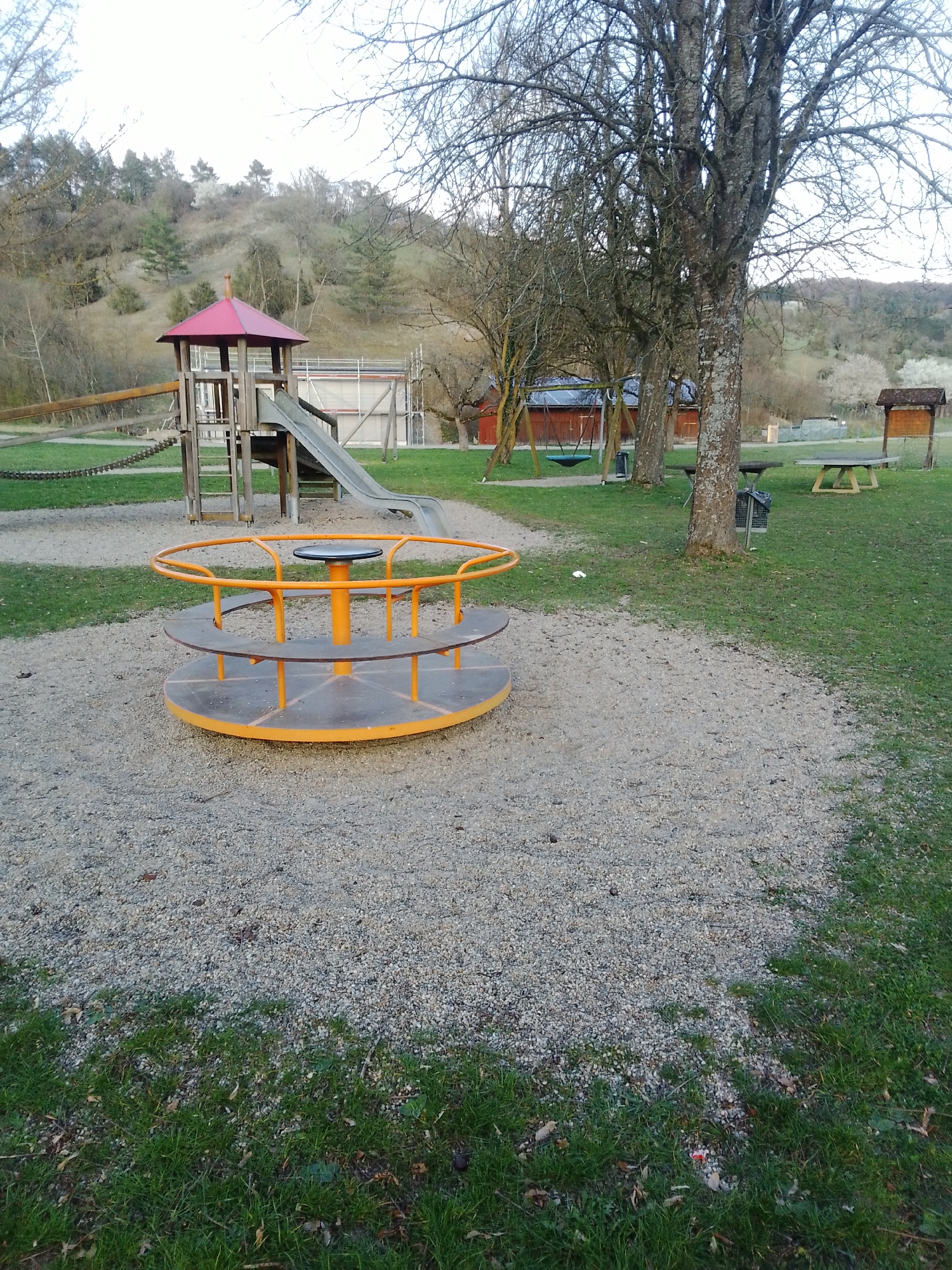 The image displays an unoccupied children's playground situated in the city of Boxberg, Germany. In the immediate foreground, a circular metal merry-go-round with orange railings and a gray platform rests on a light gray gravel surface. Green grass borders the gravel area on the left and right.
In the midground, a wooden playground structure with a red peaked roof and a gray slide is visible. A wooden walkway with chain railings extends to the left from this structure. No individuals are present on or near the playground equipment. To the right, several tall, bare deciduous trees are prominent, with a large tree trunk forming a central vertical element. Behind this tree, a swing set with a dark seat is partially visible. Further right, two wooden picnic tables and a wooden information board with a small peaked roof are situated on a grassy expanse. Two metal trash receptacles are also present near the trees.
The background features a gently sloping hillside covered with mostly bare trees and sparse vegetation, suggesting a late autumn or early spring season. To the left, several red-roofed buildings are discernible. A modern building with scaffolding is partially visible behind the main playground structure on the right. The sky is uniformly bright and overcast, casting diffuse light across the scene.