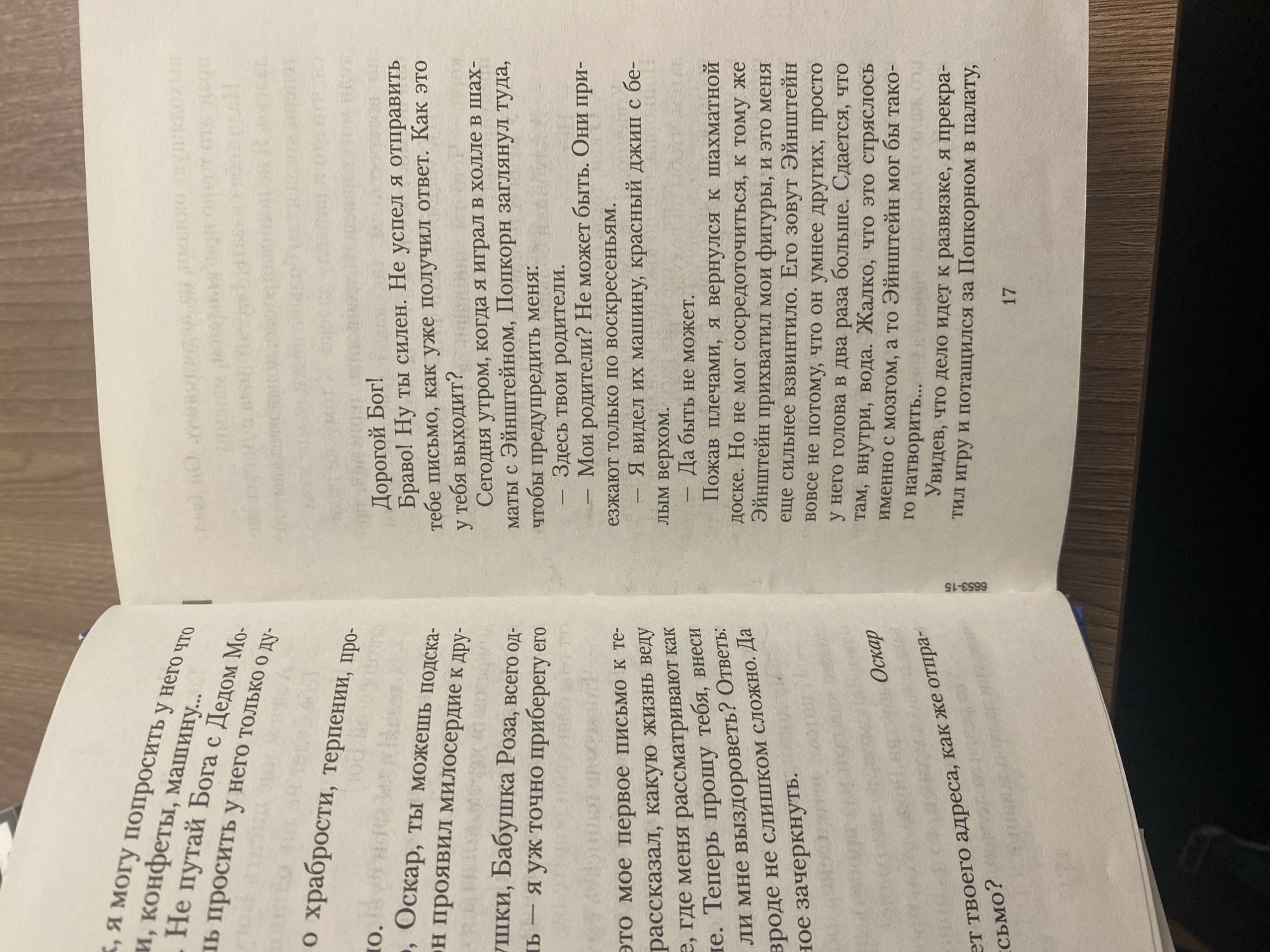 An open book, likely a paperback, is depicted resting on a flat surface, possibly a table, in what appears to be an indoor setting. The book is open to pages of text, written in Russian.
On the right-hand page, the text begins with "Дорогой Бог!" (Dear God!) and continues with a narrative featuring characters named "Эйнштейн" (Einstein) and "Попкорн" (Popcorn). The page number "17" is visible at the bottom right corner. Near the spine, a numerical code "6653-15" is partially visible.
The left-hand page also contains Russian text, mentioning "Оскар" (Oscar) and "Бабушка Роза" (Grandma Rose), and references "мое первое письмо" (my first letter). The name "Оскар" is prominently displayed at the top of this page.
The overall image suggests a moment of reading or presentation of the book's content. The implied location context for this media file is Saint Petersburg, Russia.
