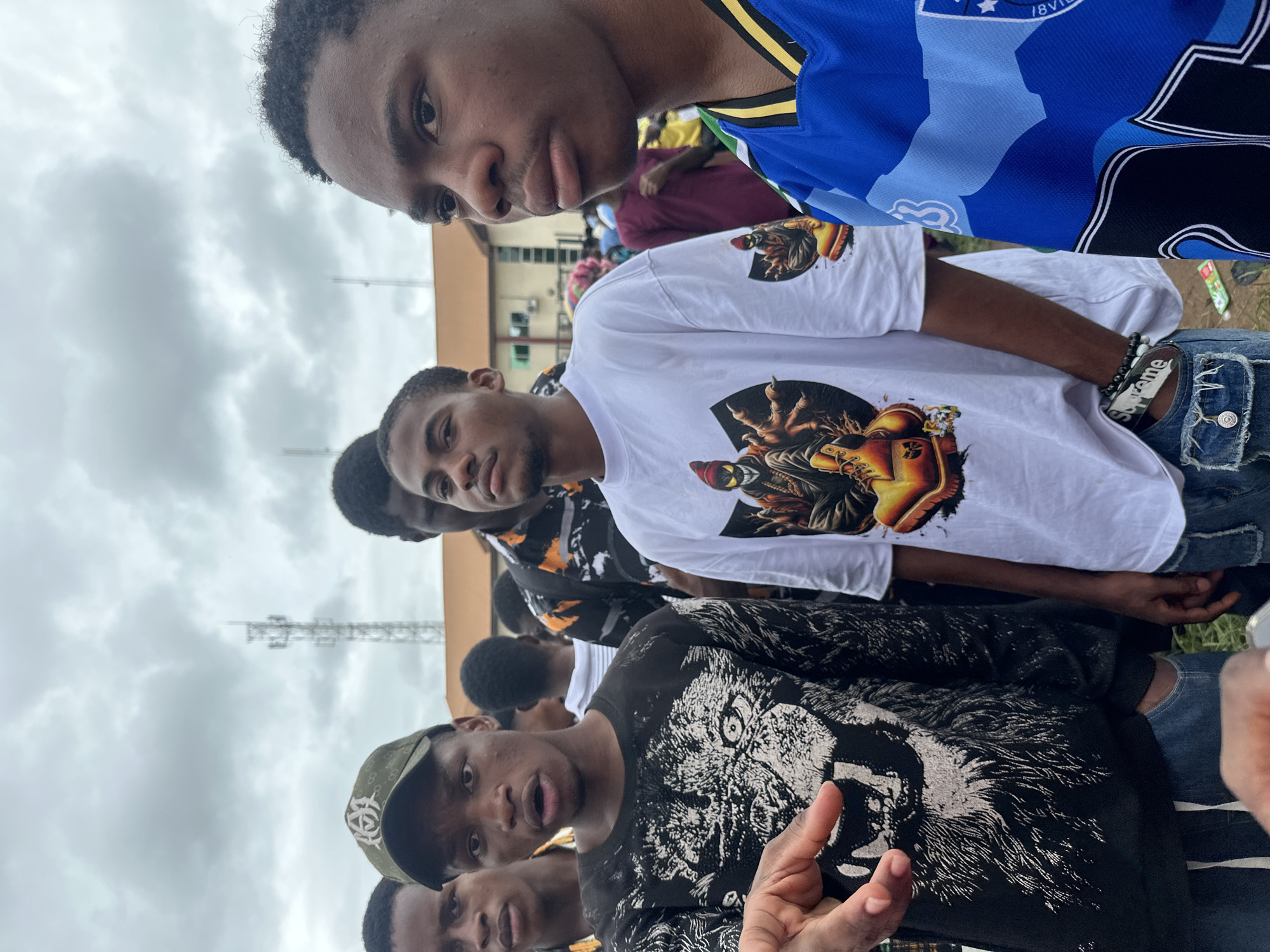 This is a medium shot photograph of a group of young Black males standing outdoors, likely in Ihiagwa, Nigeria, given the context. The sky is overcast with heavy clouds. In the background, a large cellular tower and a building with an orange roof are visible. The individuals are the primary focus, with several of them looking towards the camera or slightly off to the side.
On the left, a young male wearing a black long-sleeved shirt with a white graphic of a lion's face is looking directly at the camera with his mouth slightly open. He is wearing a green baseball cap with a logo and blue jeans. To his right, another young male in a white t-shirt with a graphic depicting a stylized character and yellow boots is looking forward. He has a bracelet on his wrist and is wearing blue jeans. To the far right, a young male in a blue and black patterned jersey with yellow trim is partially visible, looking to his right.
Other individuals are visible in the background, some with colorful shirts, suggesting a public gathering or event. The ground appears to be dirt or unpaved. The overall impression is a candid moment captured among a group of friends or attendees at an outdoor event in a Nigerian city.