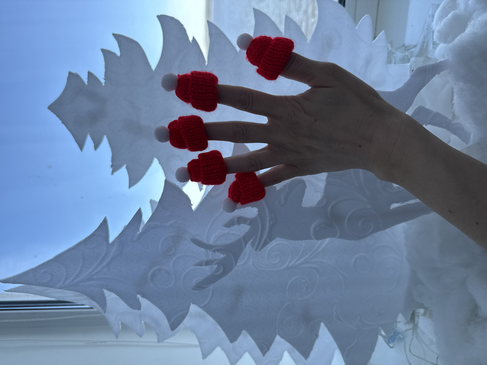 A person's hand is adorned with five small, knitted red hats with white pom-poms on the fingertips. The hand is positioned in front of a decorative white cutout of a Christmas tree, which appears to be affixed to a window. Outside the window, a blue sky is visible. The environment includes additional decorative elements, possibly cotton or a similar material, to simulate snow. The city or specific location within Votkinsk, Russia cannot be determined from the image.