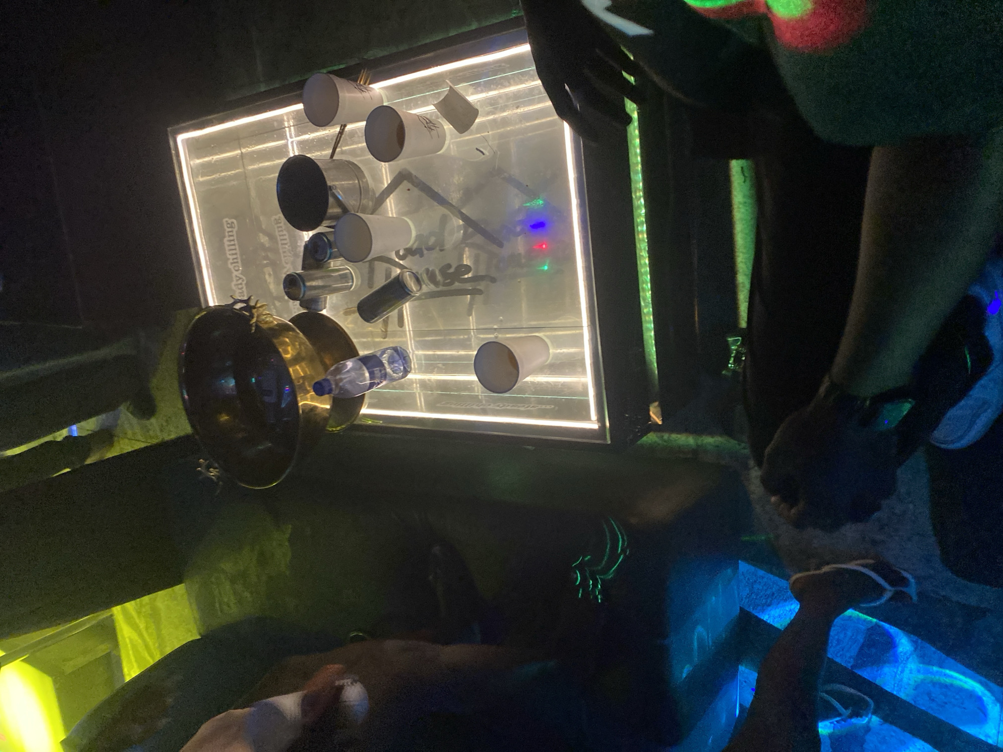 The image depicts a dimly lit scene, likely a nightclub or bar, with a focus on a table illuminated by LED lights. The table surface itself is transparent, showcasing a light-up display beneath. On the table, several white paper cups, some containing liquid, are arranged. A metal ice bucket is also present, along with a bottle of water and other miscellaneous items. In the foreground and background, parts of people are visible, including legs with flip-flops and hands. The overall ambiance is dark and atmospheric, with vibrant green and blue lighting adding to the setting. The exact location is unknown.