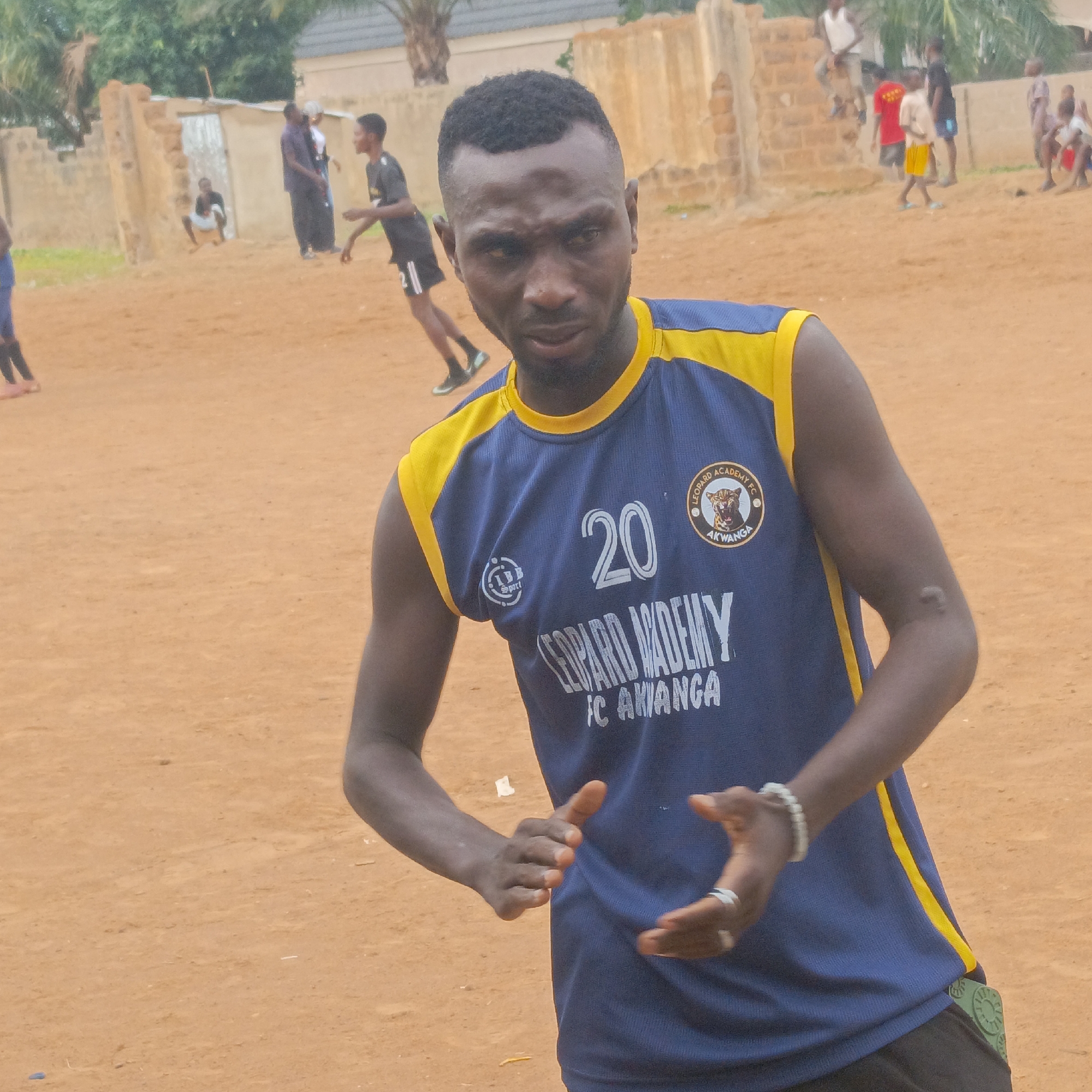 An outdoor scene depicts a group of people on an unpaved, reddish-brown dirt field, likely a sports ground. In the foreground, a dark-skinned man with a short, faded haircut and a beard looks intently at the camera. He wears a blue sleeveless sports jersey with yellow trim around the collar and armholes. On his chest, the number "20" is prominently displayed in white, along with the text "LEOPARD ACADEMY" and "FC AKWANGA." A circular patch on his right chest features a leopard's head logo and the words "LEOPARD ACADEMY FC AKWANGA." A smaller "LB Sport" logo is on his left chest. He has a white beaded bracelet on his left wrist and a silver ring on his right ring finger. His hands are slightly raised and cupped as if mid-gesture or preparing for an action.
In the background, several other individuals are present, suggesting a sports activity or gathering. On the left, a person in a black shirt is in motion, possibly running. Further back, people are standing or sitting near brick and concrete structures, with sparse vegetation and palm trees visible. One person is seated on a low wall, while others are observing or participating. The ground is bare earth, suggesting a casual playing environment. The lighting appears diffused, possibly indicating an overcast sky or twilight hours. The scene captures a moment of active engagement within a community setting in Akwanga, Nigeria.