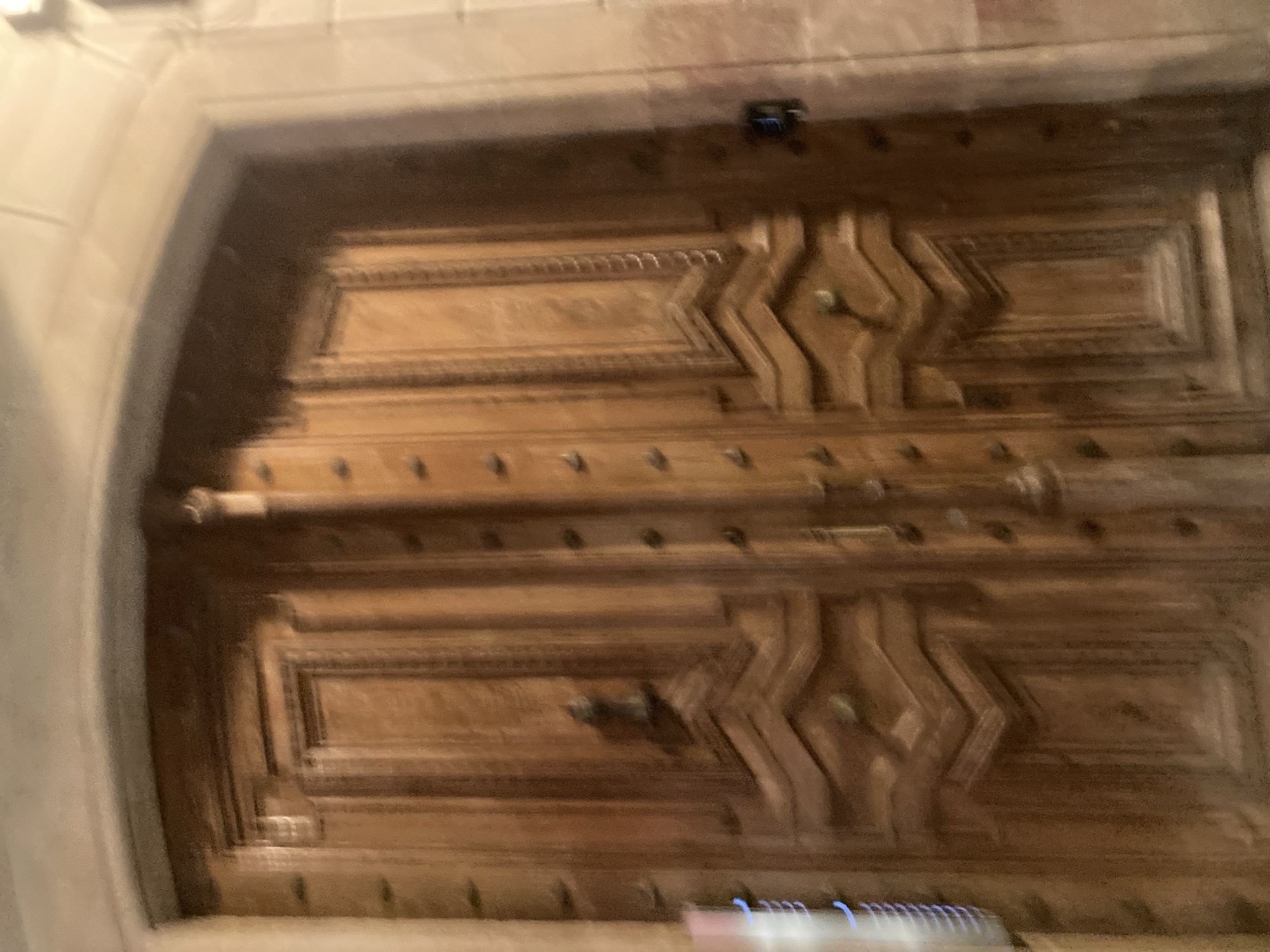 The image shows a large, ornate wooden double door, likely an entrance to a historic building. The doors are carved with intricate geometric patterns and decorative studs. A dark, metallic door knocker is visible on the left door. Above the doors, a stone archway is present. The lighting suggests it is nighttime, and the overall image is slightly blurred, possibly due to motion. On the right side of the door, a black electronic lock is attached. The architecture suggests a location in an older part of a European city.